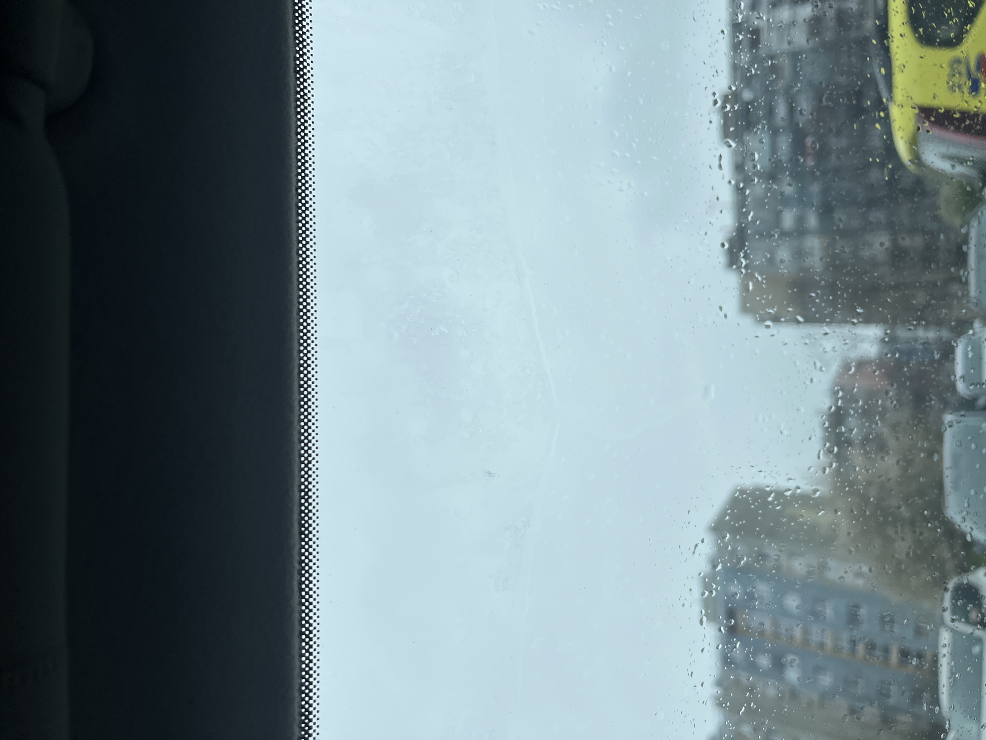 The image, taken through a rain-streaked window, shows an urban environment. A tall building is partially visible on the right side of the frame, obscured by raindrops on the glass. A yellow vehicle, possibly a taxi, is also visible on the right. The weather appears overcast and rainy. The location is likely Burgas, Bulgaria, based on available data.
