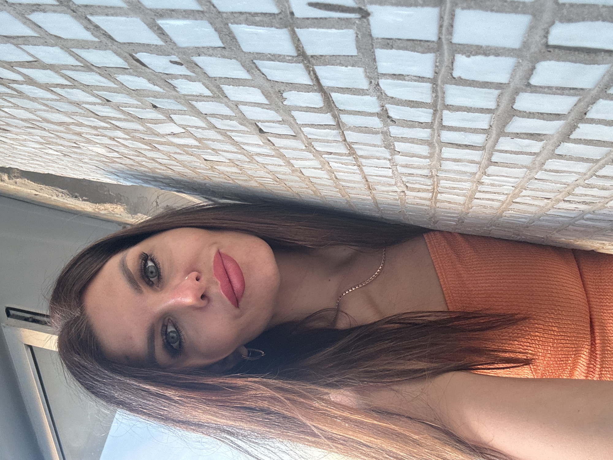 A young woman with long brown hair and green eyes is facing the camera. She has pink lipstick and a nose piercing. She wears an orange top and a gold necklace. The background shows a white tiled wall. There is no visible indication of the city of Chaikovsky, Russia.