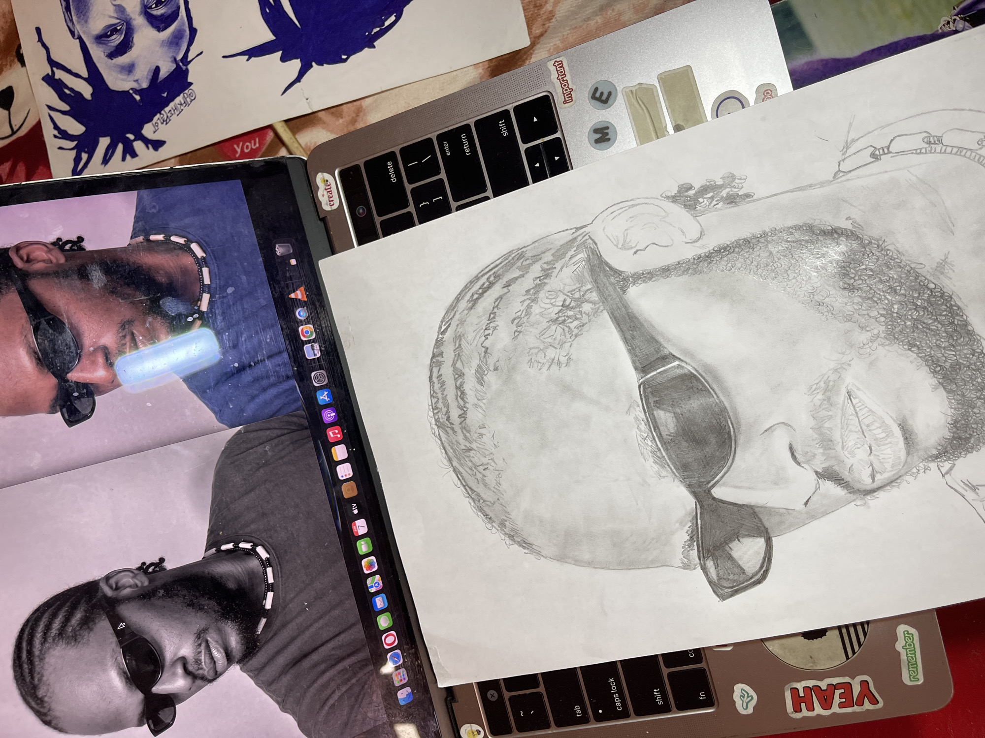 The image displays an overhead view of a workstation featuring an open silver-colored laptop, a pencil drawing, and additional sketches. The laptop screen displays two images of an individual with braided hair, sunglasses, and a beaded necklace; the left image is monochrome, while the right is in color and shows a light-emitting cylindrical object near the person's mouth. The macOS dock is visible at the bottom of the screen, along with various application icons. A hand-drawn pencil portrait of the same individual, also featuring braided hair, sunglasses, and a beard, rests on the laptop's keyboard and trackpad. Several stickers adorn the laptop's chassis, including "create," "important," "M," "E," "YEAH," and "remember." To the right of the laptop, two smaller pieces of paper contain blue ink drawings of faces with spiky hair. The background consists of a patterned fabric surface and a red surface in the bottom left corner. No visual cues within the image indicate a specific location in the city of Jos, Nigeria.
