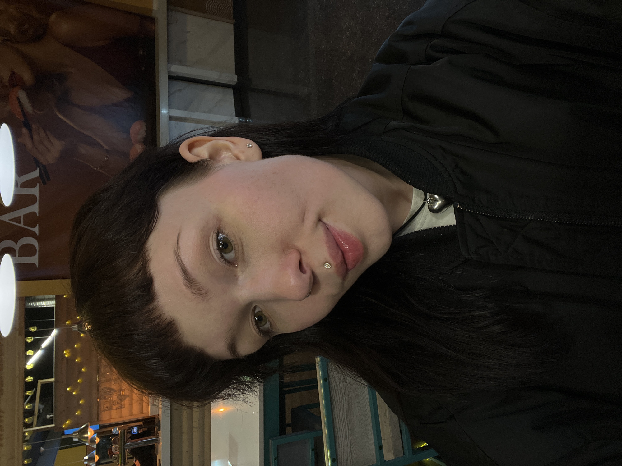 A woman with dark hair and fair skin poses in an indoor setting. She has facial piercings on her lip and ear. She is wearing a black bomber jacket. The background shows a bar with lights and signage. The location appears to be Kazan, Russia, as indicated by the Cyrillic letters on the bar sign.