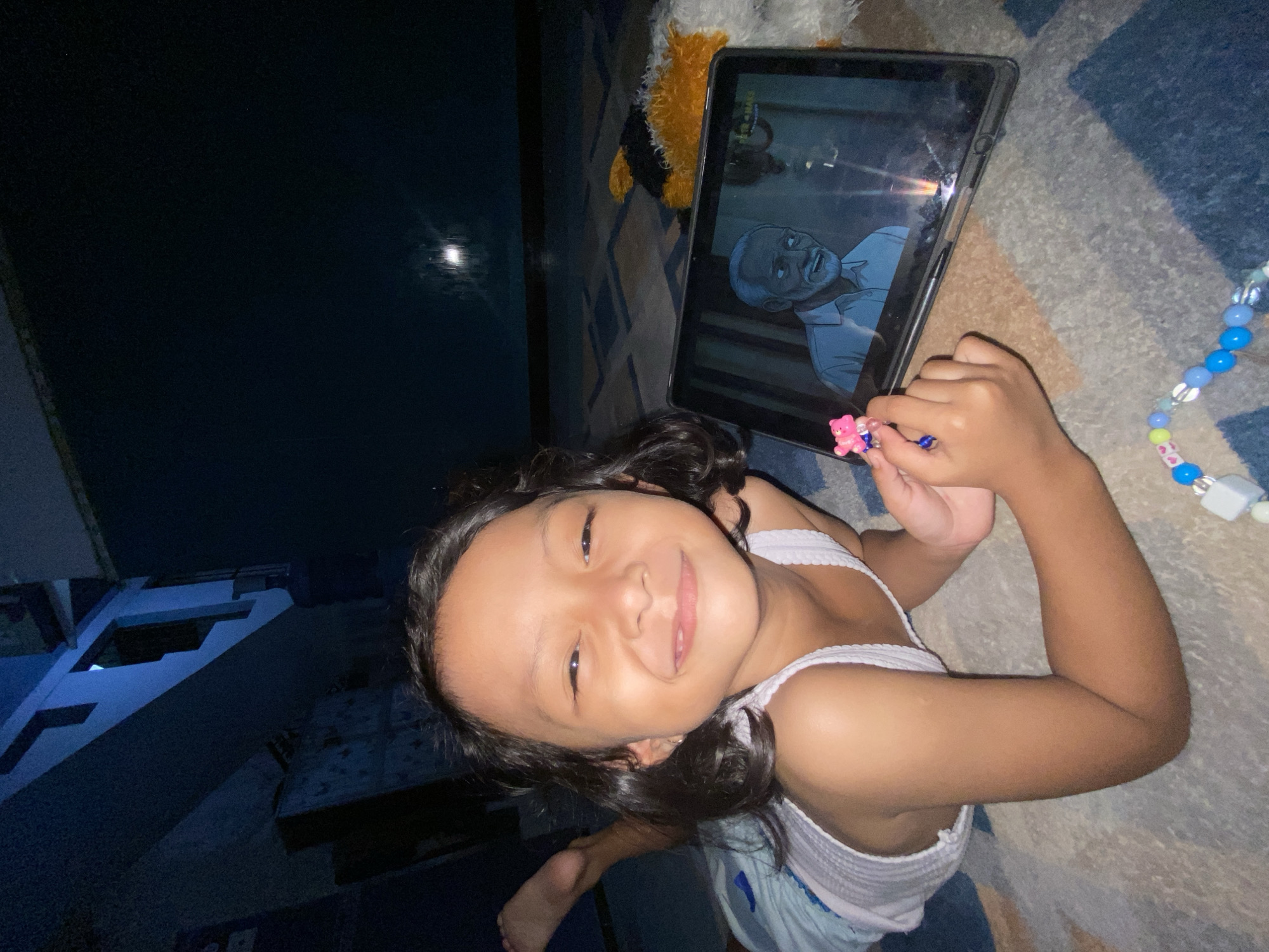 A young girl is lying on a blue and beige patterned rug, smiling at the camera. She is wearing a white tank top and what appears to be blue shorts. On her fingers are rings with small bear decorations. Beside her is a tablet displaying an animated male figure. To the left, a dark structure can be seen with blue lighting, suggesting it is nighttime in an urban area. The image was likely taken indoors, in the city of Gelam, Indonesia.