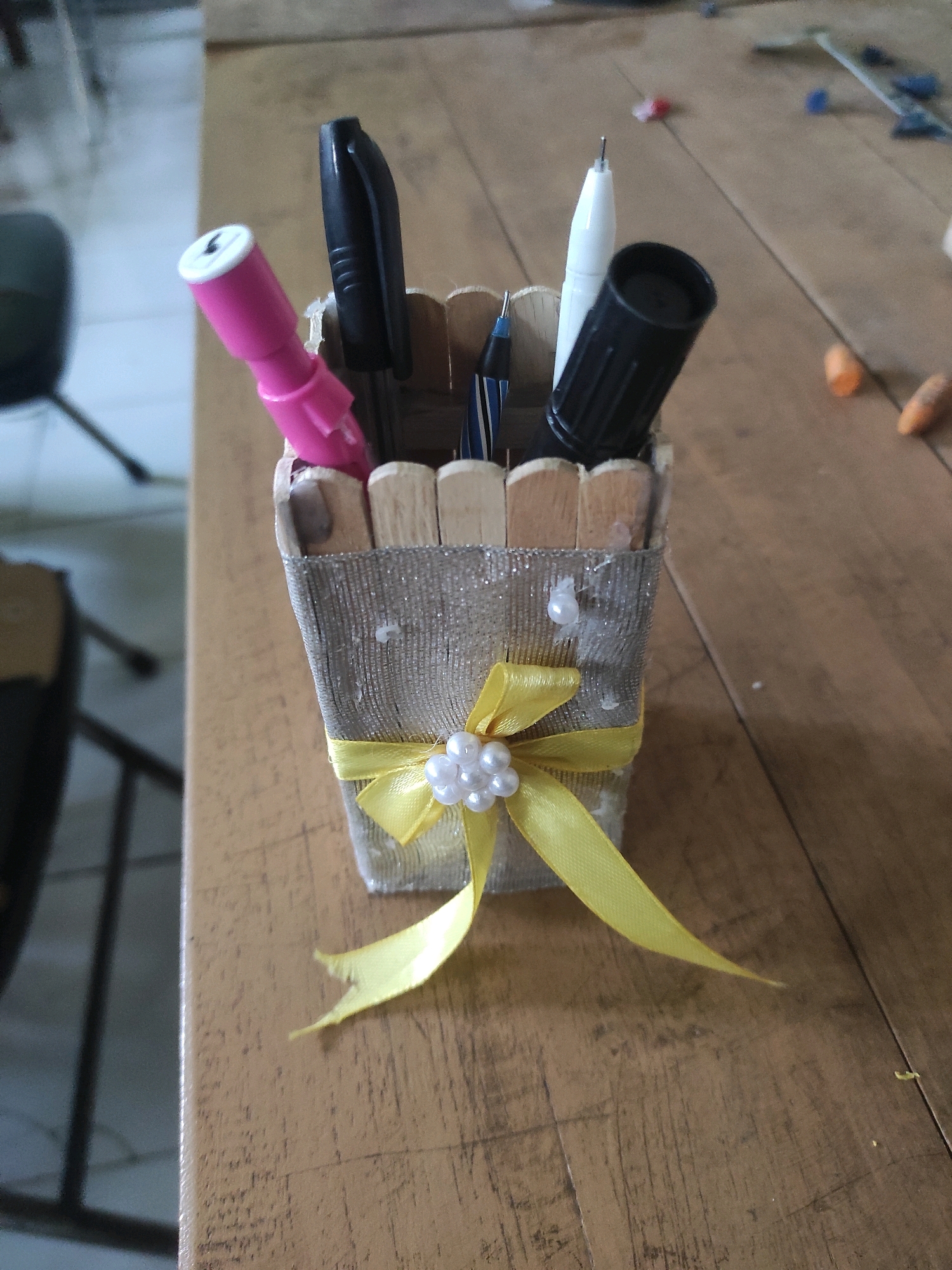 A close-up, eye-level, medium shot shows a decorated pencil holder on a wooden surface. The pencil holder is made of popsicle sticks and wrapped in a sparkly silver fabric. A yellow ribbon is tied around the front of the holder in a bow, adorned with small white beads. Several writing instruments are in the holder: a pink pen with a white tip, a black pen, a white pen, and a blue and black pen. The background is blurred, but it appears to be an indoor setting with a floor or table and some objects on it. The overall impression is that of a craft project. The specific location in Ngujung, Indonesia cannot be determined from the image.