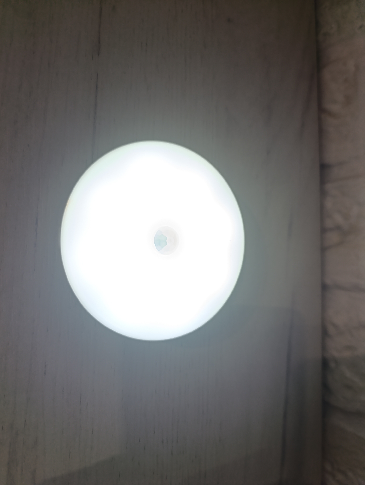 A close-up shot shows a round, white LED motion-activated night light, illuminated and emitting a bright, diffused glow. The night light is positioned on a light gray, textured surface, possibly wood paneling, with faint vertical lines. In the center of the illuminated surface of the night light, a small, recessed, multifaceted sensor is visible. The right side of the image shows a darker, rougher textured surface, possibly brick or stone. The image is taken indoors, likely in a residential setting. The scene does not depict any people, actions, or interactions.