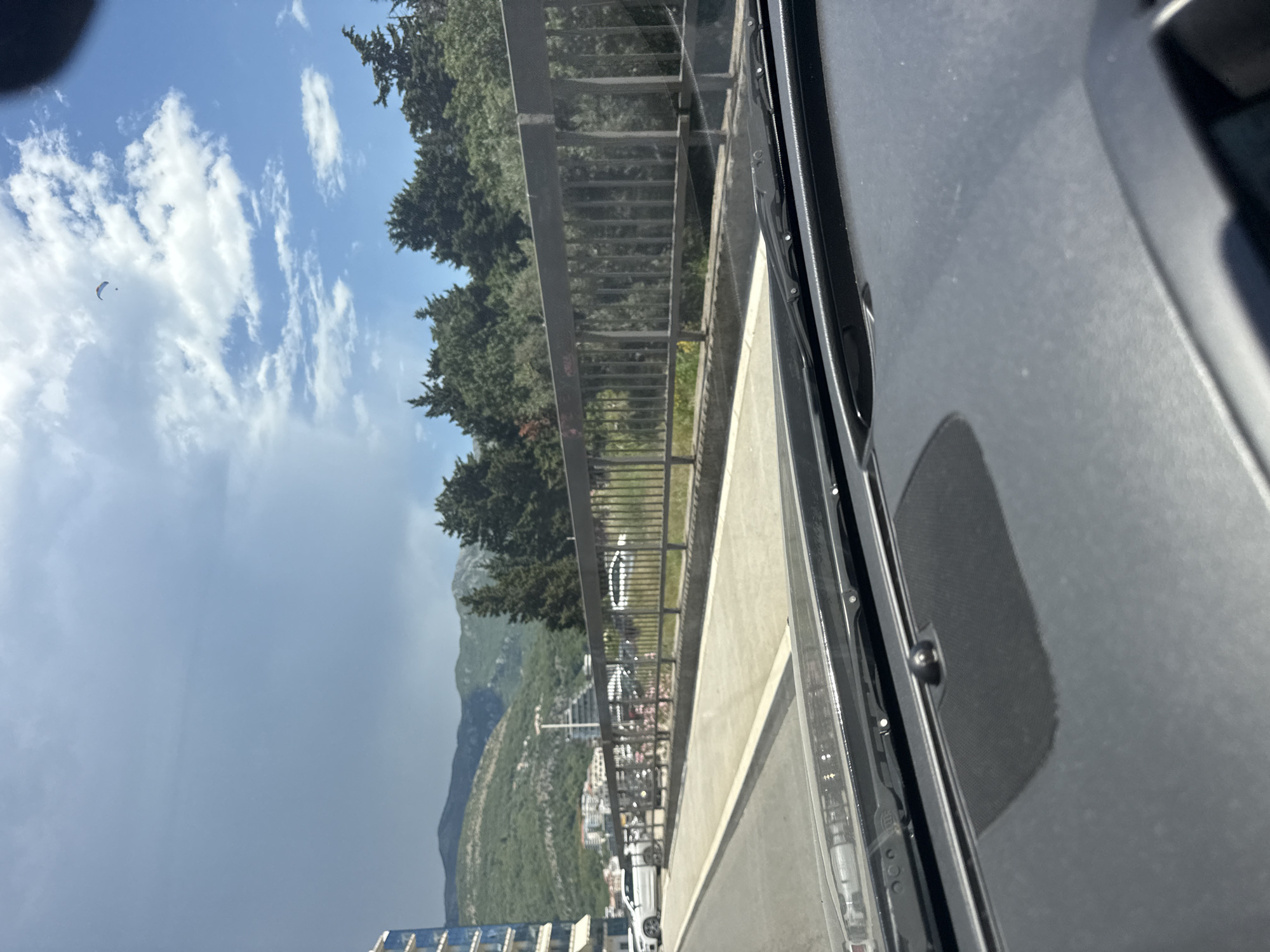 The image depicts a view from the interior of a vehicle, looking out through the windshield. The scene captures a coastal area in Boreti, Montenegro. In the foreground, the dark grey dashboard of the car is visible, with part of the windshield wipers. The road ahead is paved, with a concrete barrier and a metal railing to the right. Beyond the railing, there is a grassy area and a dense line of trees. In the background, a range of green, mountainous terrain forms the backdrop, with a scattering of buildings nestled on the slopes. The sky is partly cloudy, with a few white clouds interspersed with patches of blue. A paraglider is visible as a small, distant figure in the upper right portion of the sky. Several cars are parked along the roadside in the middle ground. A tall, blue-sided building is visible on the far left of the image.