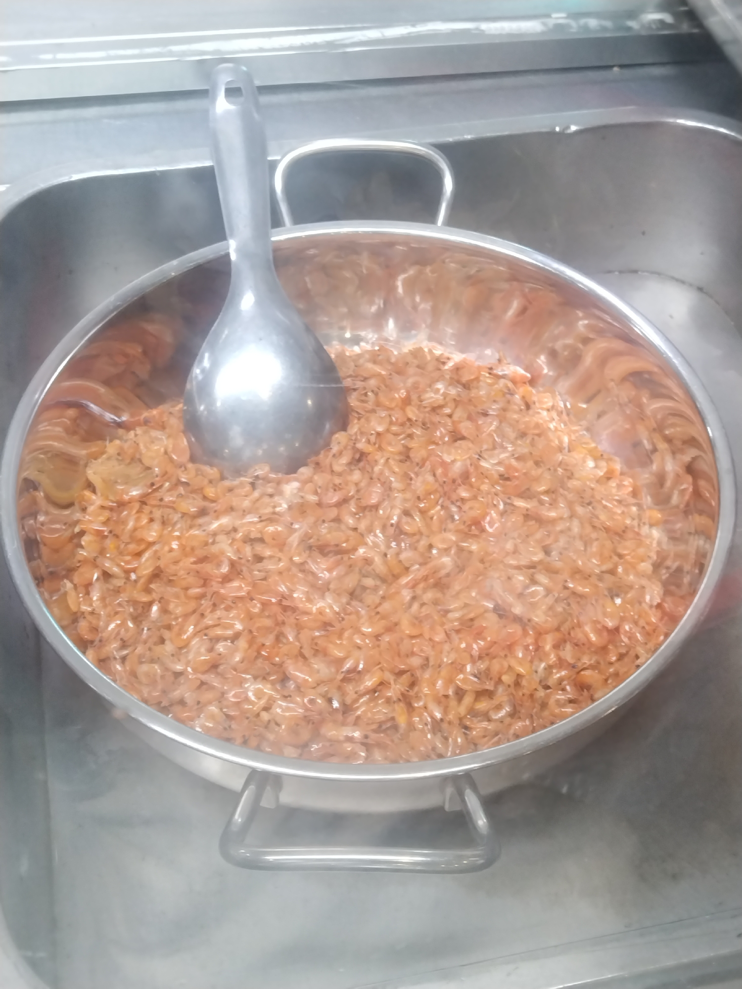 A stainless steel bowl sits within a stainless steel sink. The bowl is filled with small, orange shrimp, with a stainless steel ladle resting in the shrimp. The scene is located within Chu Văn An Ward, Vietnam.