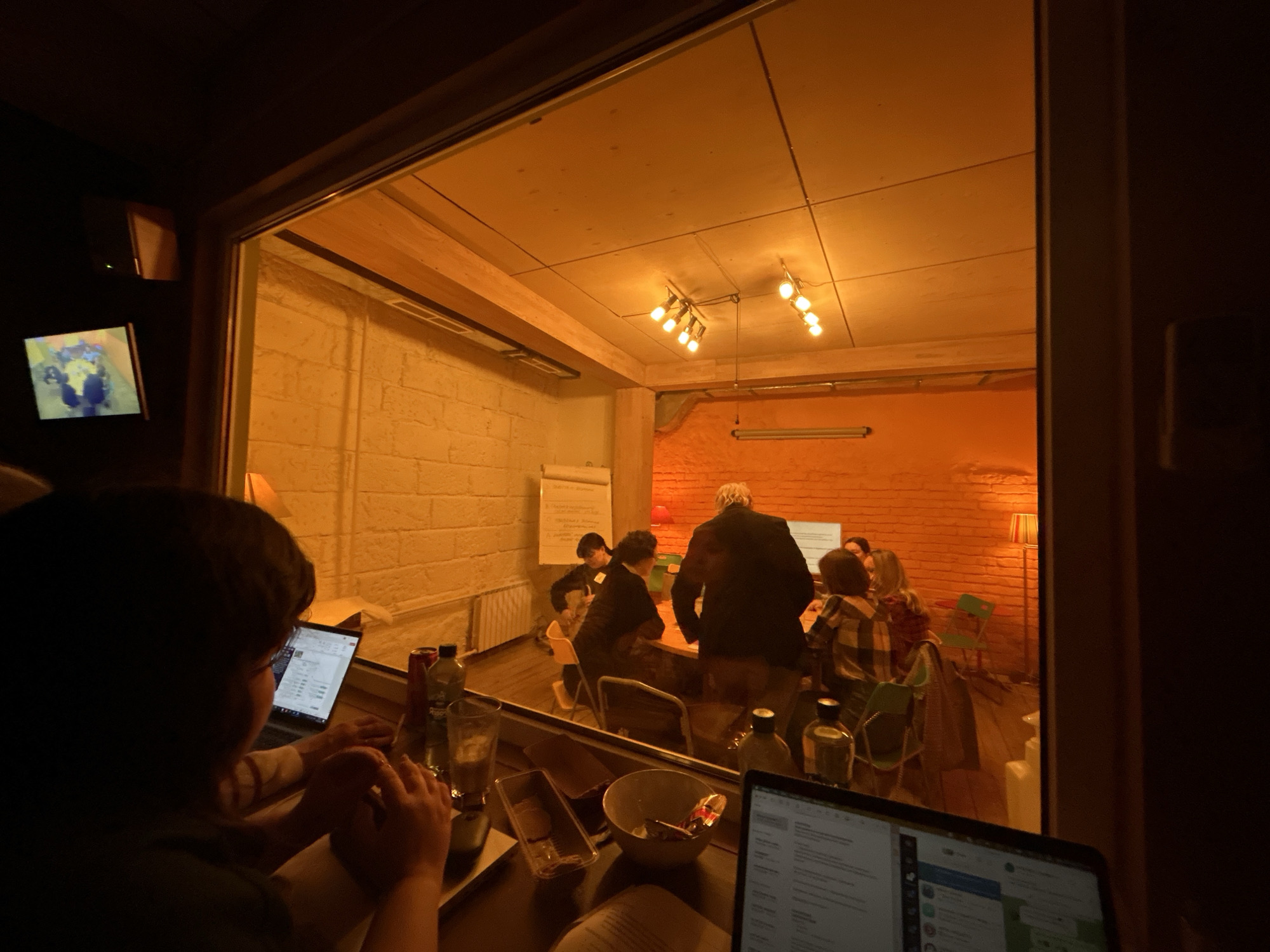 The image shows an interior scene, likely a meeting or work space, viewed from an adjacent darker room or area. A group of people is gathered around a table in a brightly lit room. A white board with notes or a presentation on it is visible. A person with a light-colored shirt in the foreground is working on a laptop. The overall lighting suggests a warm or yellow tint. The location appears to be a collaborative or co-working space. The image was captured in Moscow, Russia.