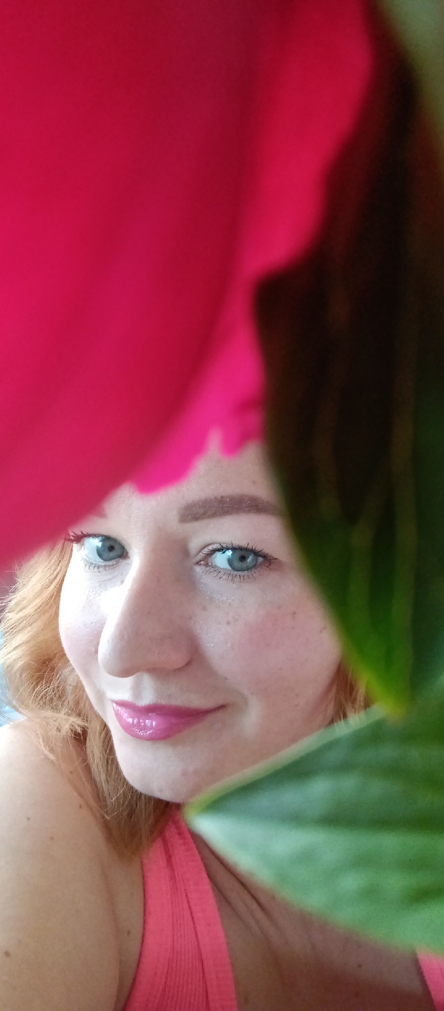 A person with fair skin, red hair, and blue eyes poses closely with a pink flower on the left and a green leaf on the right, partially obscuring the view of their face and body. The person is wearing pink lipstick and a pink top. Freckles are visible on their face. The location and environment are unknown.
