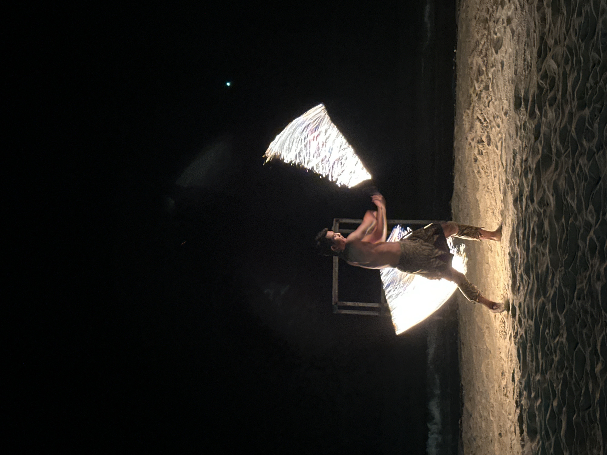 The image shows a shirtless male standing on a sandy beach, presumably at night in Hat Khai Muk, Thailand. He is performing a fire dance with two triangular torches. The beach is dark, but the sand is illuminated by the fire. In the background, there is a dark structure. A small, bright point of light is visible in the upper left. The image is rotated 90 degrees clockwise.
