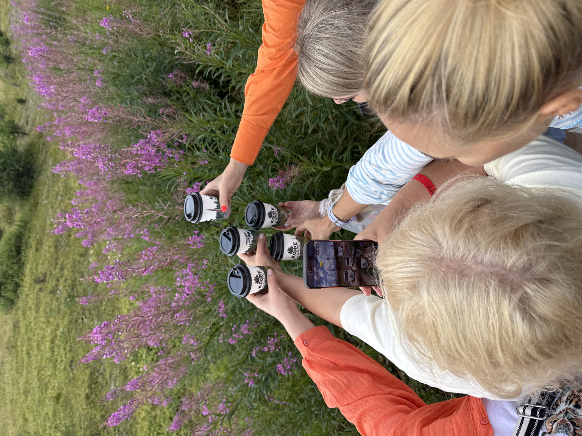 In an outdoor setting with a background of green grass and purple flowering plants, several individuals are gathered. One person is holding a smartphone, taking a photograph or video of the scene. Several hands are extended, each holding a small, black-lidded coffee cup with white branding. The cups appear to be for takeaway beverages. The attire suggests a casual gathering, with visible clothing including an orange shirt and a blue and white striped shirt. The overall impression is a moment captured during an outing in a natural environment. The specific location of T/B Seminskii Pereval, Russia, is not discernible from the visual elements of the image itself.