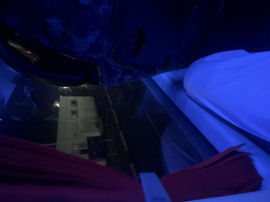 The image displays a nighttime scene observed from inside a vehicle, located in Starovoitove, Ukraine. The interior of the vehicle is illuminated by a strong blue ambient light. On the left, a pleated red curtain is drawn back by a light-colored plastic or fabric tie. In the lower center, the legs of an individual, clad in white fabric, are visible, prominently illuminated by the blue light. To the right, a dark blue patterned seatback, featuring a light-colored ornamental design, is partially visible. Through the window, a dark exterior environment is present. A multi-story, light-colored building with three visible square windows is dimly illuminated in the mid-ground. In front of the building, a paved surface is visible, exhibiting horizontal light-colored markings and a dark, pole-mounted object or sign. No individuals are distinctly visible outside the vehicle, and no overt actions or interactions are depicted.
