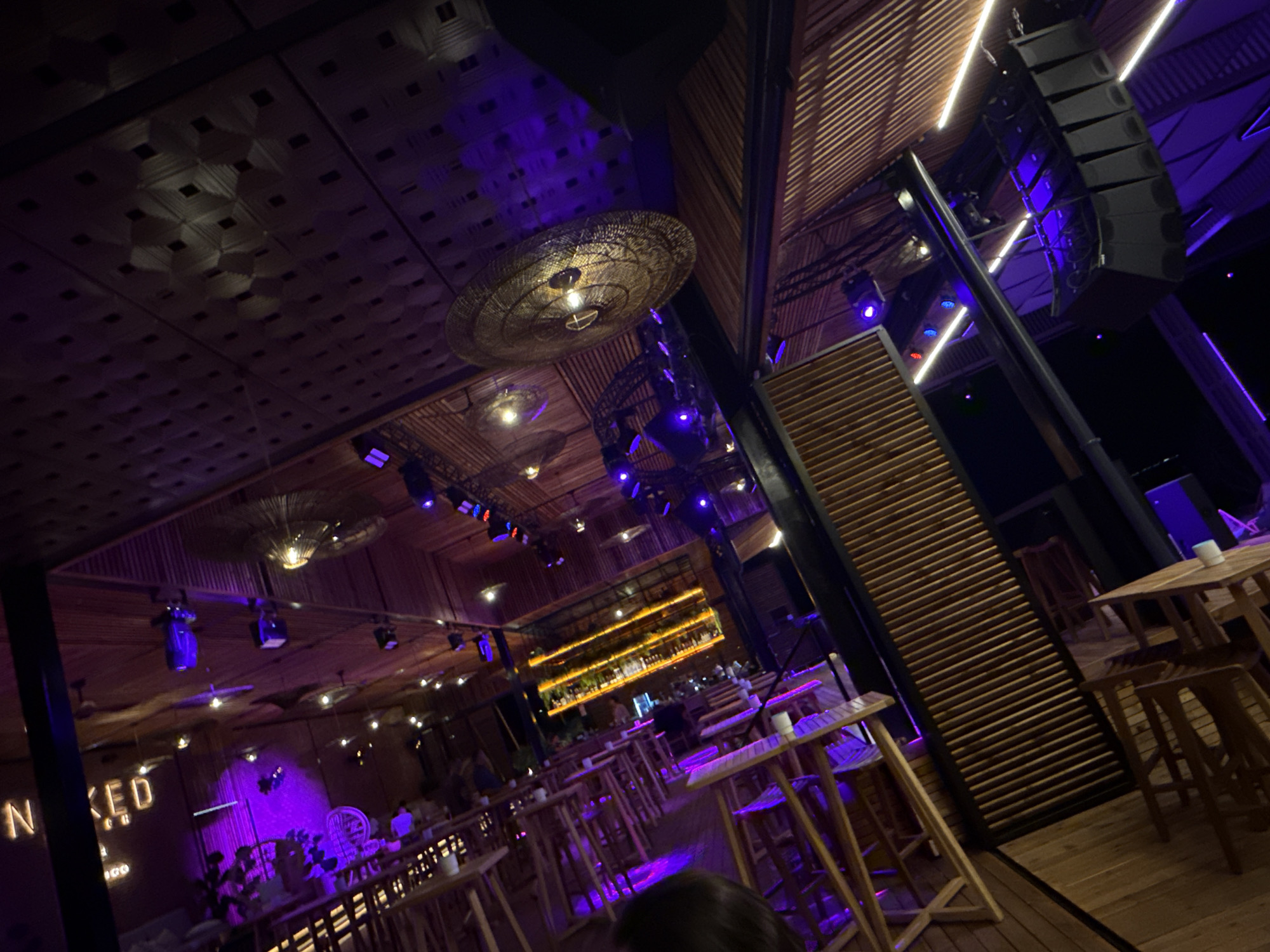 This media file depicts the interior of a modern, dimly lit establishment, likely a bar or club, in Burgas, Bulgaria. The scene is set at night, illuminated by a combination of purple and white stage lighting, creating a vibrant and energetic atmosphere. The architecture features a rustic aesthetic with wooden ceilings and walls, adorned with decorative lamps and an impressive array of speakers mounted on truss systems, suggesting a venue that hosts live music or DJ sets.
The primary subjects are the sparse patrons scattered throughout the space. A few individuals are visible at tables, some appearing to be socializing, while others are more solitary. The lighting casts a purple hue over much of the scene, bathing the tables and seating in a cool, atmospheric glow. The arrangement of tables and chairs indicates a space designed for both dining and entertainment, with high-top tables and bar stools visible.
Notable details include the distinctive ceiling design, a patterned wooden structure that catches the light, and the prominent speaker system, indicating a focus on sound and performance. A bar area is visible in the background, illuminated by a warm, golden light from rows of bottles and shelving, contrasting with the dominant purple lighting. There is also a sign visible that partially reads "NAKED" and "GOOD," possibly part of the venue's branding or a call to action. The overall impression is one of a lively, stylish nighttime venue ready for patrons to enjoy music and drinks.