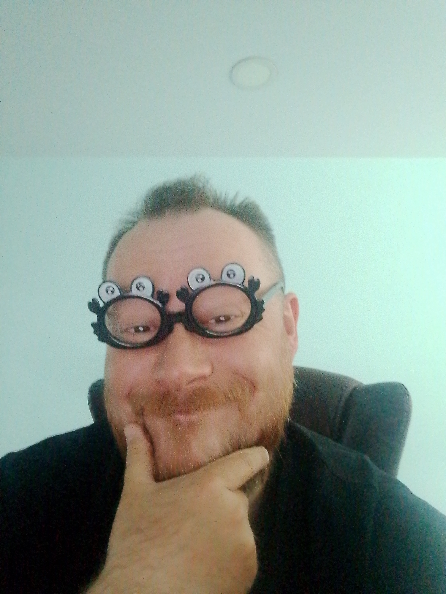 A selfie shows a fair-skinned male with a red beard and mustache wearing novelty glasses with large cartoon eyes. He is seated, appearing from the shoulders up, wearing a dark shirt. His right hand is raised with his index finger touching his chin in a pensive gesture. The background is a light blue wall with a white ceiling and a recessed circular light fixture. The location is not discernible from the image.