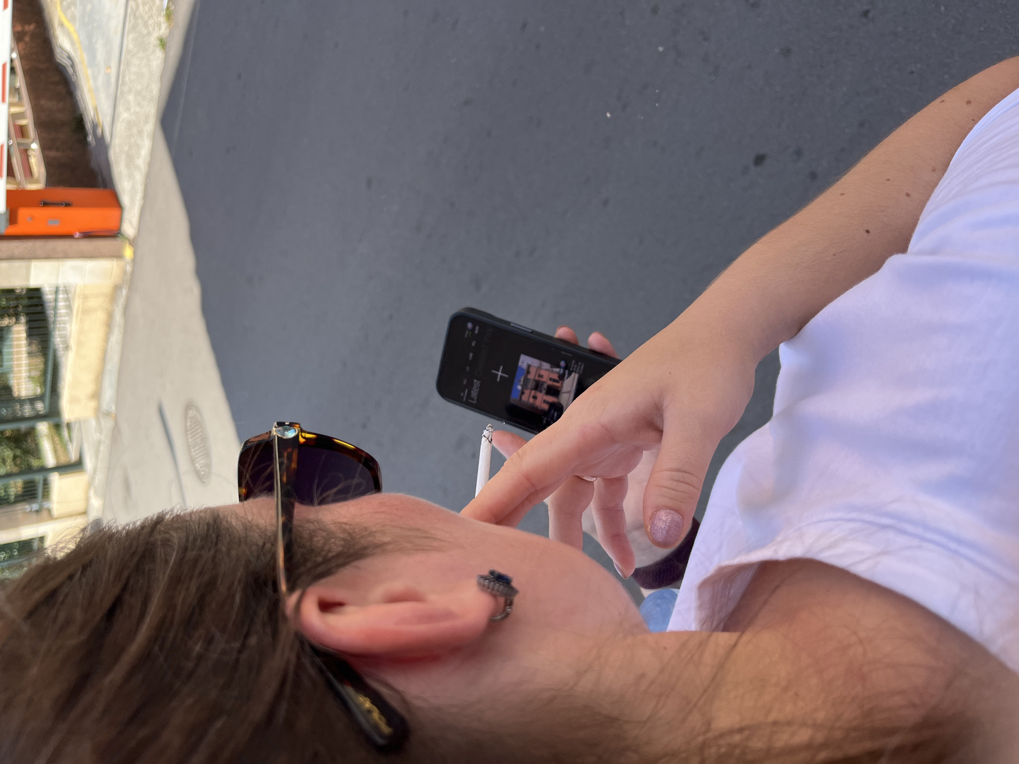 The image shows a close-up, high-angle shot of a person in Budva, Montenegro. The person is wearing sunglasses and has a cigarette in their mouth. They are holding a smartphone in their hand, which displays a musical interface with the word "Latest" and a plus sign. The person is also wearing an earring. Their arm, visible in the frame, is lightly tanned and has some moles. They are wearing a white t-shirt.
The scene appears to be outdoors, with a street or sidewalk visible in the background, suggesting an urban or semi-urban environment. The lighting indicates it is daytime, likely sunny. The overall mood is casual and relaxed. No specific time of day is discernible beyond it being daylight. No weather cues are present other than the bright lighting.