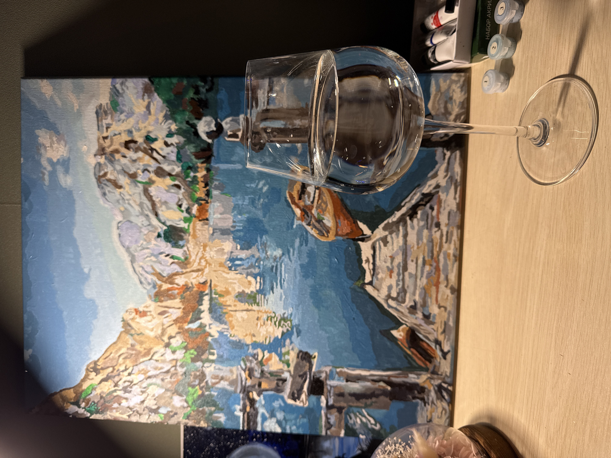 The image depicts a still life arrangement on a wooden surface. In the foreground, a wine glass filled with clear liquid is prominently featured. Behind the glass, a painting of a landscape is visible, showing mountains reflecting in a body of water. A small wooden dock extends into the water, with a boat tied to it. To the right of the glass, several small containers of paint and a box labeled "НАБОР АКРИЛ" (Acrylic Set in Russian) are arranged. To the left, a glass dome with dried flowers is partially visible. The lighting suggests an indoor setting, possibly a desk or table. The scene is likely a personal workspace or display. The image does not contain any recognizable landmarks or elements that would allow for a specific location in Cheboksary, Russia, to be identified.