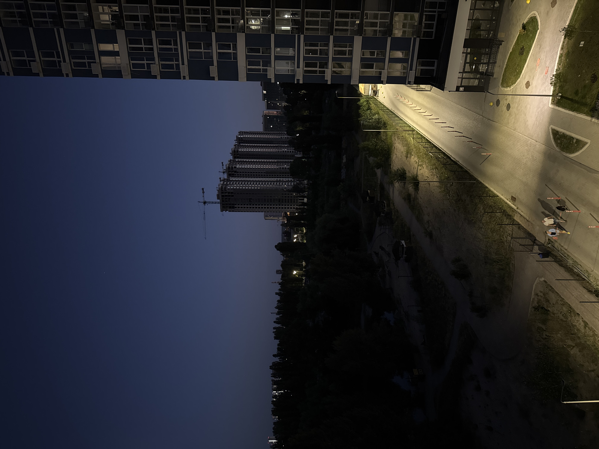 A nocturnal, high-angle shot captures an urban landscape under a deep blue twilight sky. To the right, a tall, multi-story building with a dark blue facade dominates the foreground. Its windows, some illuminated from within, create a pattern of light against the dark exterior. Below the building, a section of a paved area with grass islands and yellow bollards is visible.
The middle ground reveals a paved pathway with lampposts casting pools of light onto the surface. The pathway is lined with a fence and orange and white bollards. Two individuals, one wearing a white shirt and shorts and the other in a dark top and lighter shorts, walk along this path. A small dog walks beside them. Further into the scene, a strip of dark green trees forms a dense, shadowy barrier across the landscape.
In the background, several tall, unfinished high-rise buildings under construction are silhouetted against the sky, with cranes visible above them. A cluster of distant city lights twinkle on the horizon, suggesting more urban development beyond the immediate area. The overall scene depicts a developing urban environment at dusk, with a mix of completed and under-construction structures, a park-like area, and people moving through the illuminated pathways.