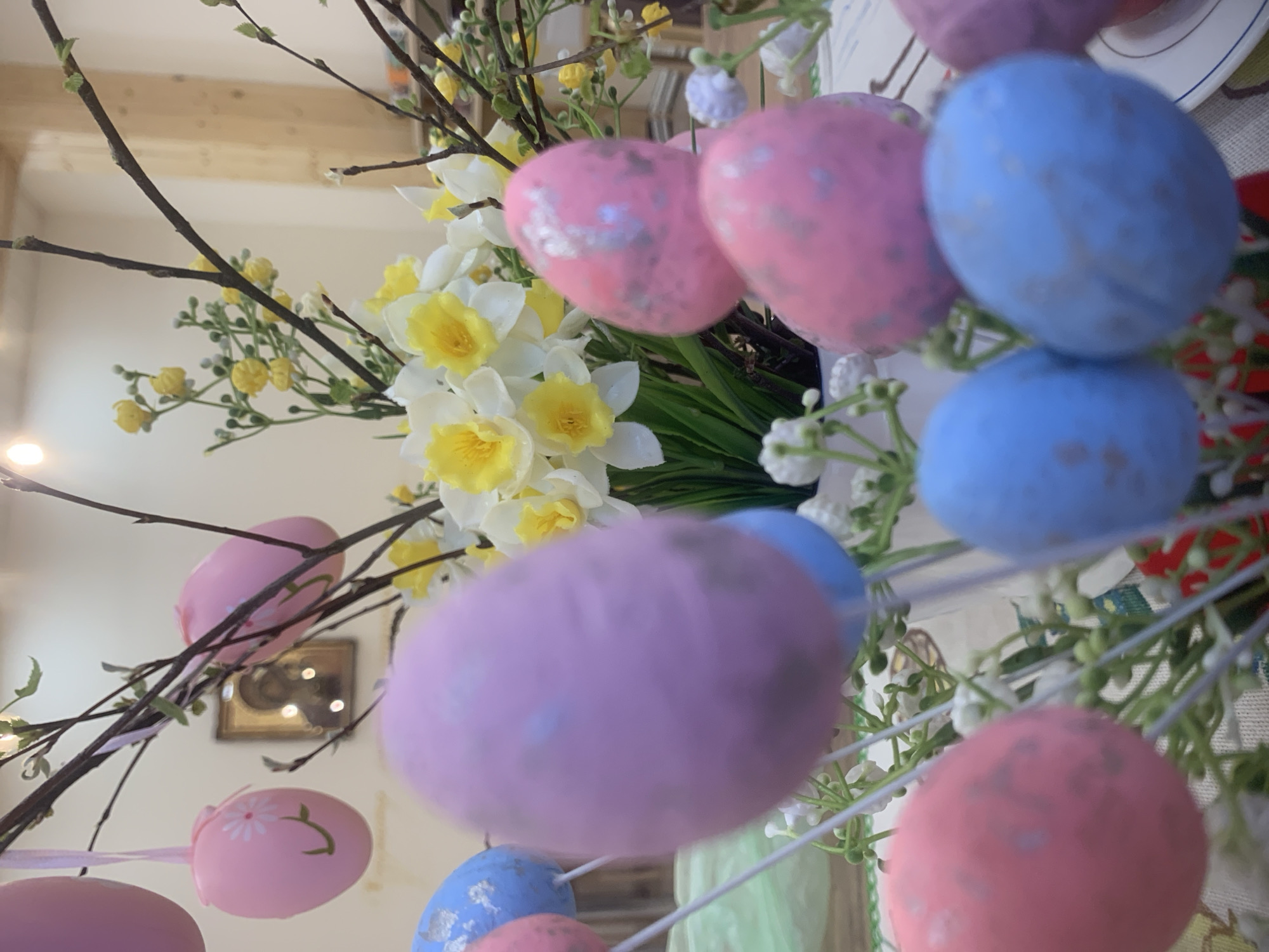 The image shows a close-up of an Easter decoration. There are pink and blue painted Easter eggs hanging from branches, intermingled with yellow and white daffodils and small white flowers. The background is blurry, but it appears to be an indoor setting with a wooden beam visible. A framed picture hangs on the wall in the background. There is no evidence to indicate that the location is Izhevsk, Russia.