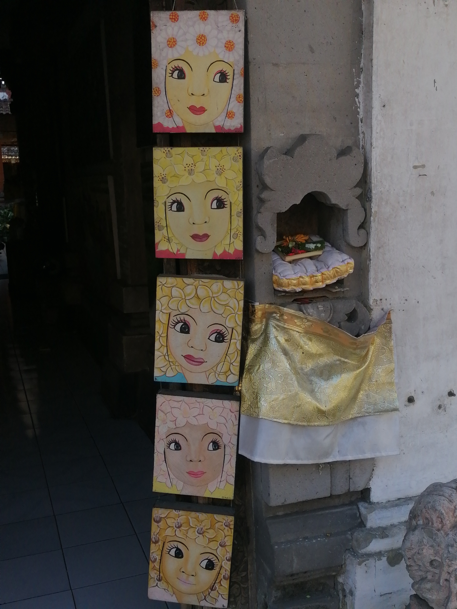 The image depicts a vertical arrangement of five small, square paintings of stylized female faces, each adorned with flower-like headpieces, hung on a rough, gray stone wall. The paintings are spaced evenly, one above the other. To the right of the paintings, a small, recessed alcove is visible in the wall, containing a cushion with gold trim and what appears to be offerings. Below the alcove, a draped piece of gold and white fabric hangs, partially obscuring the wall and a stack of rough stones at the base. A weathered, carved stone figure is visible on the far right of the frame. The background to the left shows a dark, shadowed doorway or entryway with tiled flooring. The style of the paintings and the presence of the stone carving and alcove suggest a location in Bali, possibly a temple or a traditional dwelling in the Ubud District, Indonesia. The scene appears to be outdoors, under natural sunlight, given the shadows and highlights.