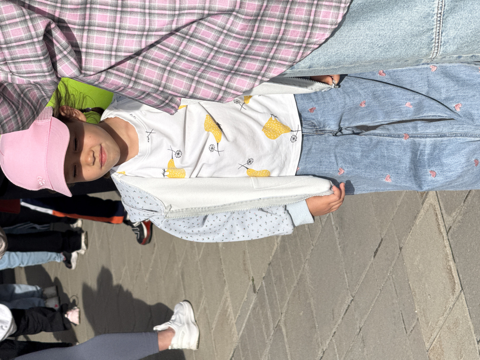 A young girl wearing a pink baseball cap, a white T-shirt with a bird pattern, and blue jeans with small heart patterns is standing on a paved surface. A light blue jacket hangs loosely around her shoulders. The girl is positioned in a group, with legs and shoes of other individuals visible around her. A pink and gray plaid shirt is draped above her. The setting appears to be an outdoor area with paving stones. There is no information from the image to indicate that the location is Yekaterinburg, Russia.