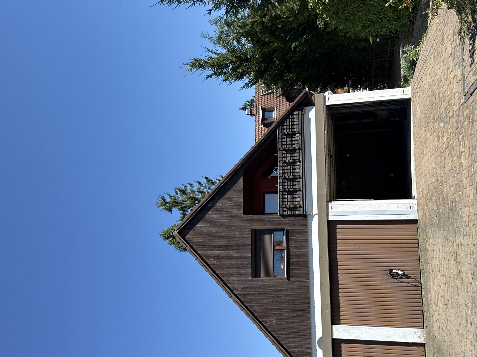 The image shows the exterior of a residential building with an attached garage in Bad Sachsa, Germany. The building has a dark brown wooden facade on the upper level, with a pitched roof covered in brown tiles. There is a window with a dark roller shutter on the upper facade, and a balcony with a wooden railing above the garage. The garage has three sections, two of which have closed brown doors. The middle section has an open garage door, revealing a dark interior. A coiled black and white cable is attached to the wall of the rightmost closed garage door. Evergreen trees and bushes are visible around the property, and the ground in front of the garage is paved with interlocking bricks. The sky is clear and blue.