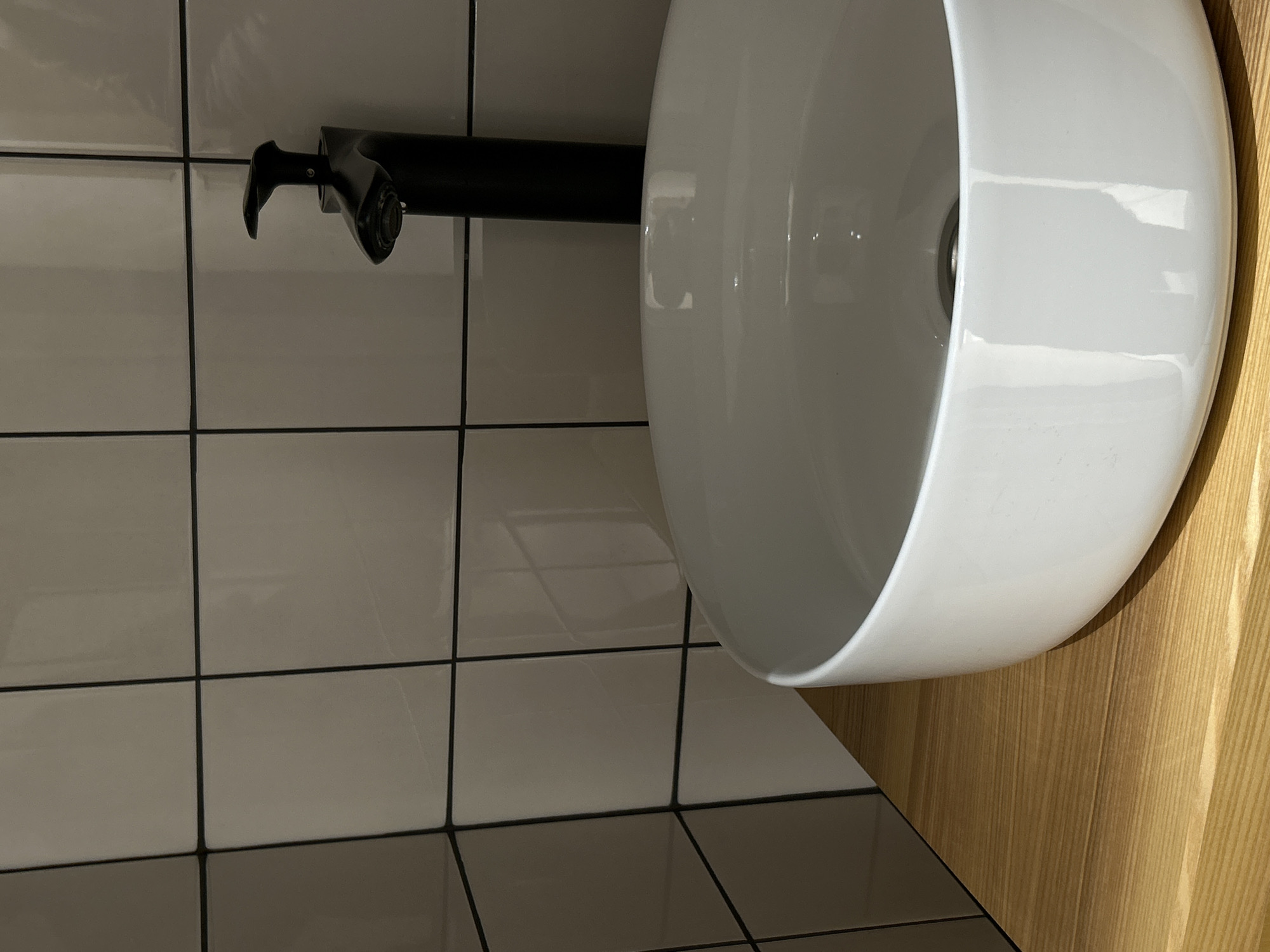 The image shows a close-up of a modern bathroom sink with a black faucet. The sink is white and has a smooth, rounded shape. The background consists of white square tiles with black grout lines. A wooden countertop is visible on the right side of the image. There are no people visible in the image. The location is not identifiable as being in Saint Petersburg, Russia from the available details.