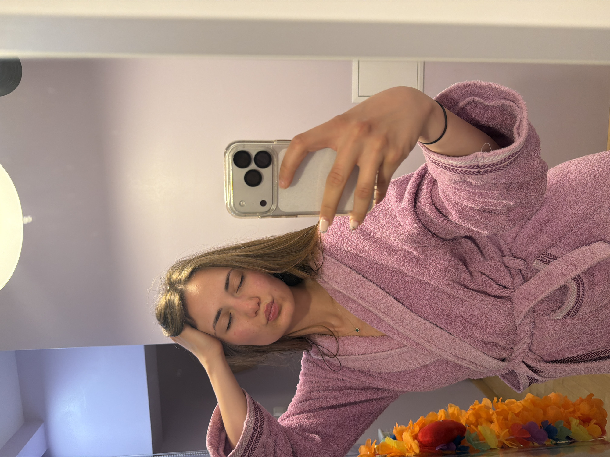 A young woman is captured in a mirror selfie in an indoor setting, likely in Buhovo, Bulgaria. She is wearing a soft pink terrycloth bathrobe with decorative striped trim on the cuffs and a tie around the waist. Her long, light brown hair frames her face.
The woman is holding a smartphone with a clear case and a triple-camera system, positioning it to take the mirror selfie. Her eyes are closed, and her lips are slightly pouted, conveying a playful, relaxed, or perhaps mock-tired expression. She rests her left hand gently on her head, while a black elastic band is visible on her right wrist. A delicate necklace with a small green pendant is visible around her neck.
The background, seen in the mirror's reflection, consists of a light-colored wall, possibly a soft lavender or pale pink. A white light fixture is partially visible in the upper left, and a white electrical switch or outlet is seen on the wall in the upper right. In the foreground, below the woman's reflection, there is a vibrant floral garland primarily composed of orange and yellow flowers, with some red and blue elements, resting on a surface. The room is brightly and evenly lit. No visible text is present in the image.