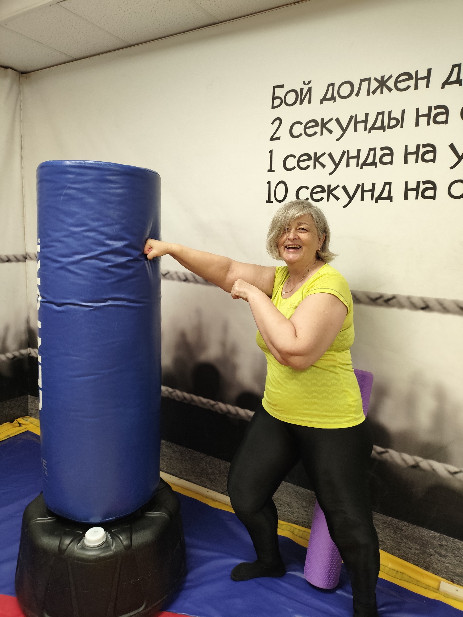A smiling, middle-aged woman with short, gray hair, wearing a bright yellow t-shirt and black leggings, stands in front of a large blue punching bag in a gym. Her right fist is positioned as if she is about to punch the bag. The gym has a wall with text in Russian, which appears to be workout instructions, and a background image of a boxing ring. There is a purple yoga mat visible behind her. The floor is a blue and red mat. This scene is located in the city of Krasnodar, Russia.