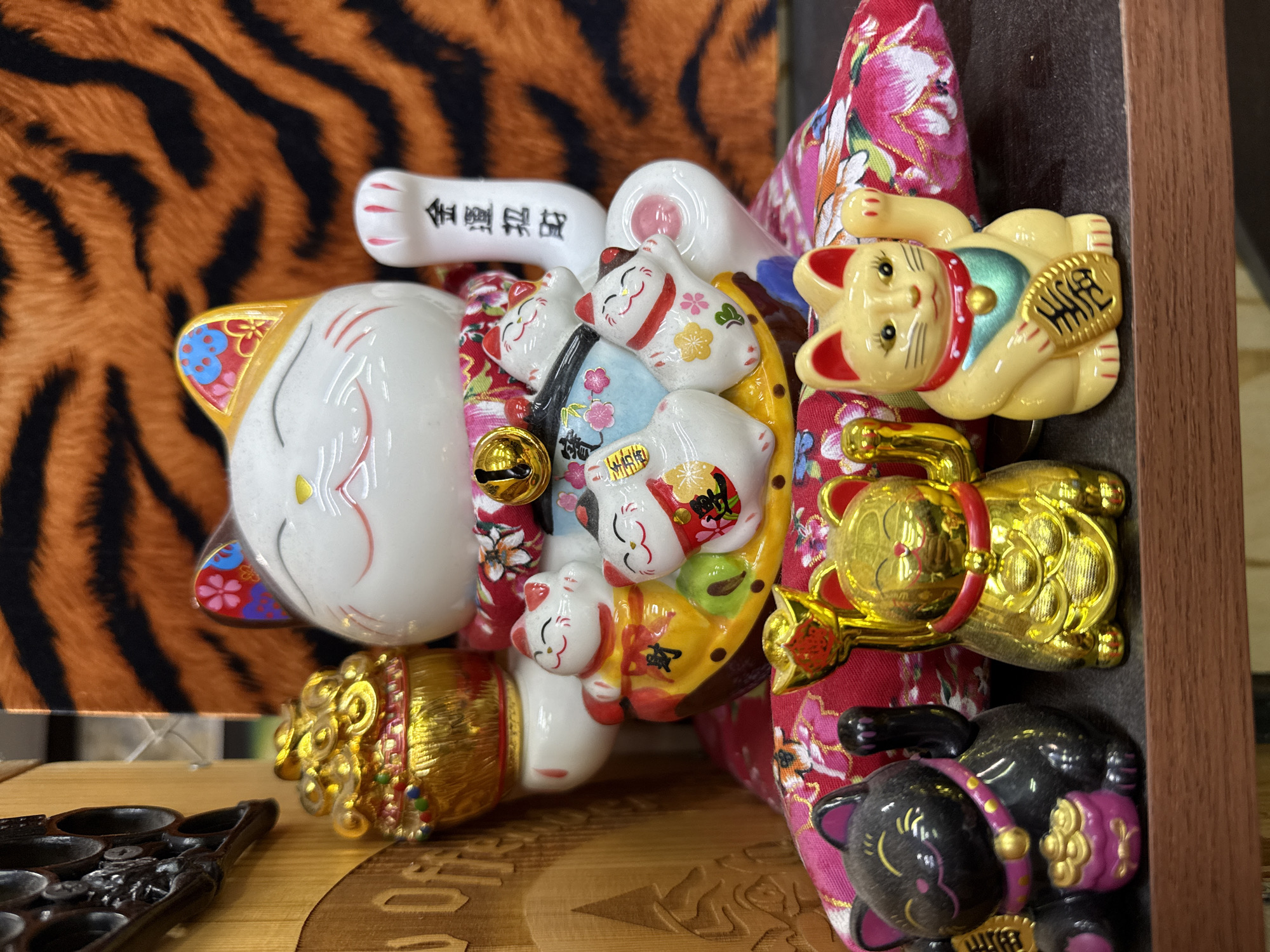 The image displays a vibrant collection of Japanese Maneki-neko (beckoning cat) figurines, primarily white, yellow, and black, arranged in a visually rich setting. The dominant figure is a large white Maneki-neko, smiling, with decorative red markings on its face and ears, and colorful floral patterns (blue, red, gold) on its side. Its raised right paw features black Japanese kanji characters, "金運来福" (Kin'un Raifuku), meaning "Good fortune and happiness come." Its belly is adorned with multiple smaller, white Maneki-neko figures, some wearing patterned kimonos (red, blue, yellow) and holding golden coins or ingots. A small golden bell hangs from the large cat's neck.
To the right, two other prominent Maneki-neko figures are visible against a backdrop of red fabric with pink and green floral patterns. The upper right shows a smaller, yellow Maneki-neko with red accents and a blue bib, holding a golden oval coin with black characters typically signifying wealth (likely "千万両" - ten million ryo). Below it, a black Maneki-neko with pink ears and collar also holds a similar golden coin, reinforcing themes of prosperity.
Numerous golden elements, including ingots and coins, are scattered throughout the display. The background features a striking orange and black tiger-stripe pattern. In the bottom-left foreground, a dark, intricately carved wooden object, possibly a decorative tray or game board, rests on a lighter wooden surface. The scene appears to be a curated display, likely in a shop or home, showcasing these traditional good luck charms. No specific activity or time of day is discernible.