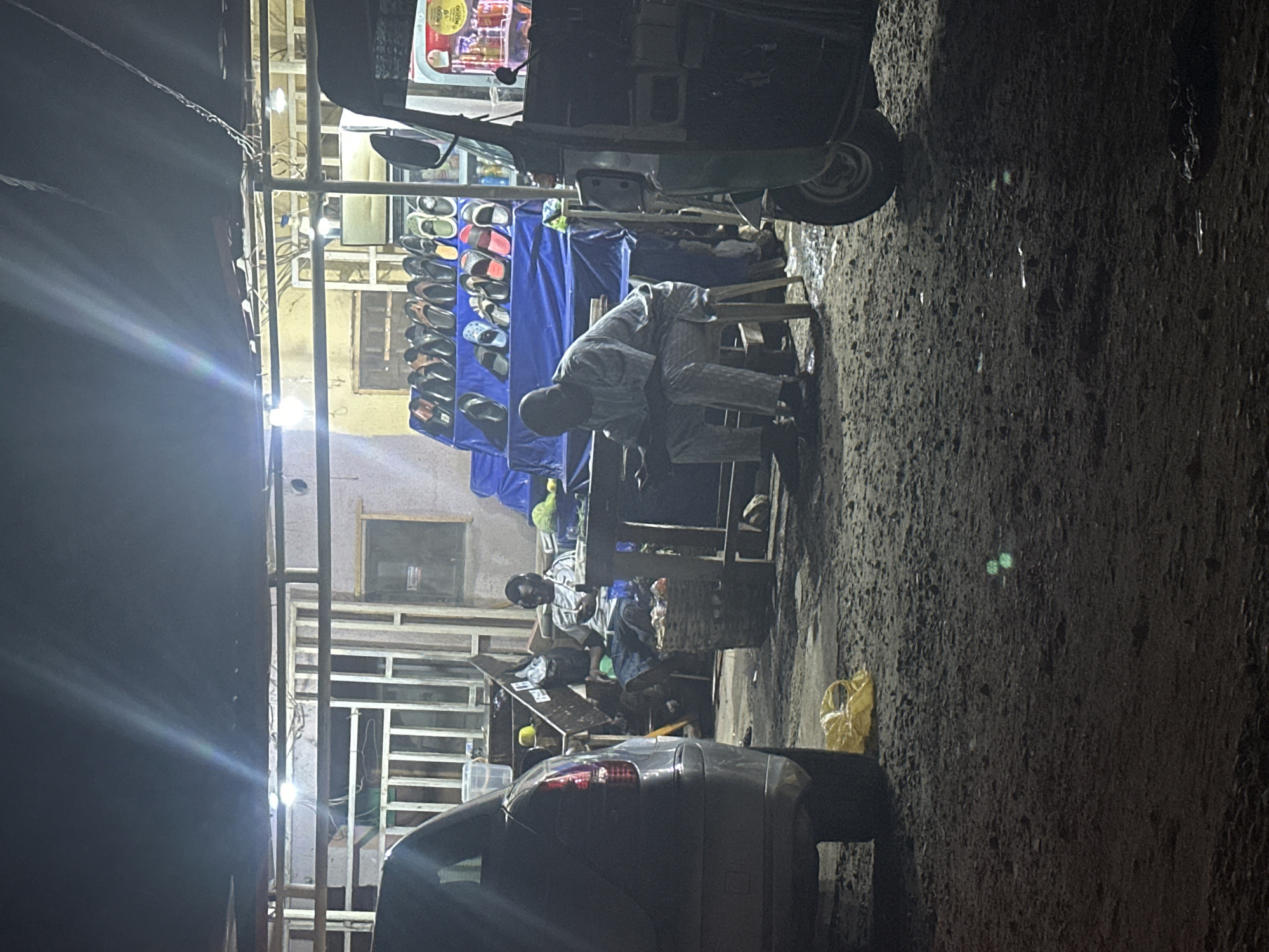 This is a night-time photograph taken in Abuja, Nigeria, depicting an outdoor market scene. In the foreground, a dark-colored car is partially visible on the left, with a yellow plastic bag on the ground in front of it. To the right, a green and black auto-rickshaw is parked. In the mid-ground, a man wearing a light-colored patterned robe is seated on a stool, facing a table. He appears to be looking down. Behind him and to the left, another man wearing a grey and white striped shirt and blue shorts is seated on a basket, looking at his phone and wearing headphones.
The background features a market stall with a blue cloth covering a display of various footwear, including sandals and flip-flops. Lights are illuminating the area, creating bright streaks in the dark sky. A metal structure with beams and a roof covers part of the market. The ground is a rough, gravelly surface. Several other objects, including crates, bags, and possibly more merchandise, are scattered around the market area. The overall impression is one of a busy, albeit dimly lit, nocturnal marketplace.