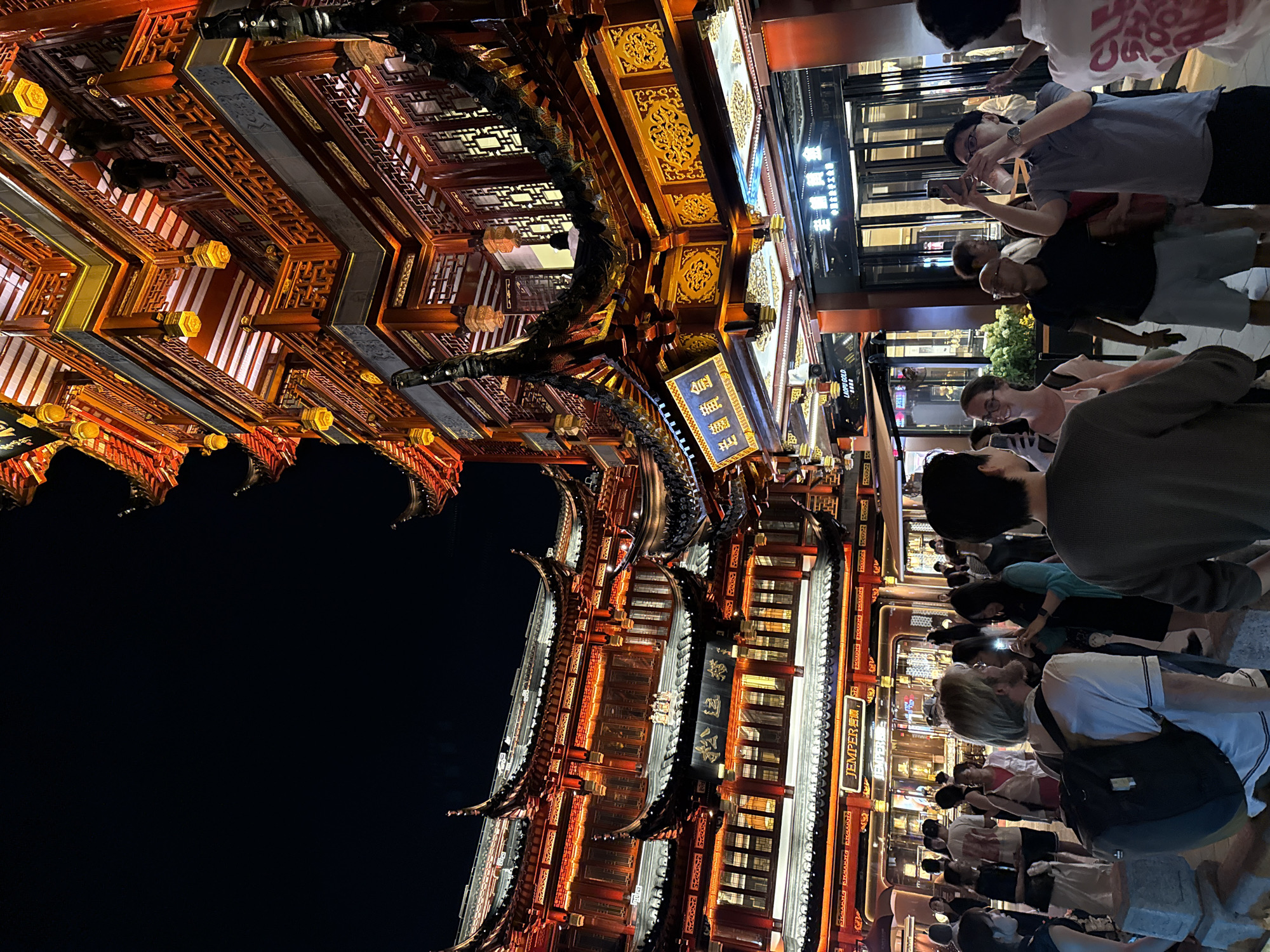 The image depicts the Yu Garden Bazaar in Shanghai, China, at night. The architecture is traditional Chinese, with multi-tiered roofs, red and gold ornamentation, and illuminated lanterns. The bazaar is crowded with people browsing shops, taking photos, and socializing.
In the foreground, a man with blonde hair and a beard, wearing a white t-shirt and a backpack, is looking to his right. A woman with glasses, wearing a light-colored top, is to his left, also looking to her right and holding a phone, possibly taking a photo. To the right of them, a man in a grey polo shirt is holding up a phone and taking a photo, while another man in a dark shirt stands behind him. More people are visible in the mid-ground and background, some walking, some standing in groups, and some entering shops. The shops appear to be selling various goods, with illuminated signage. The overall atmosphere is lively and bustling, illuminated by the warm glow of the traditional architecture and shop lights against the dark night sky.