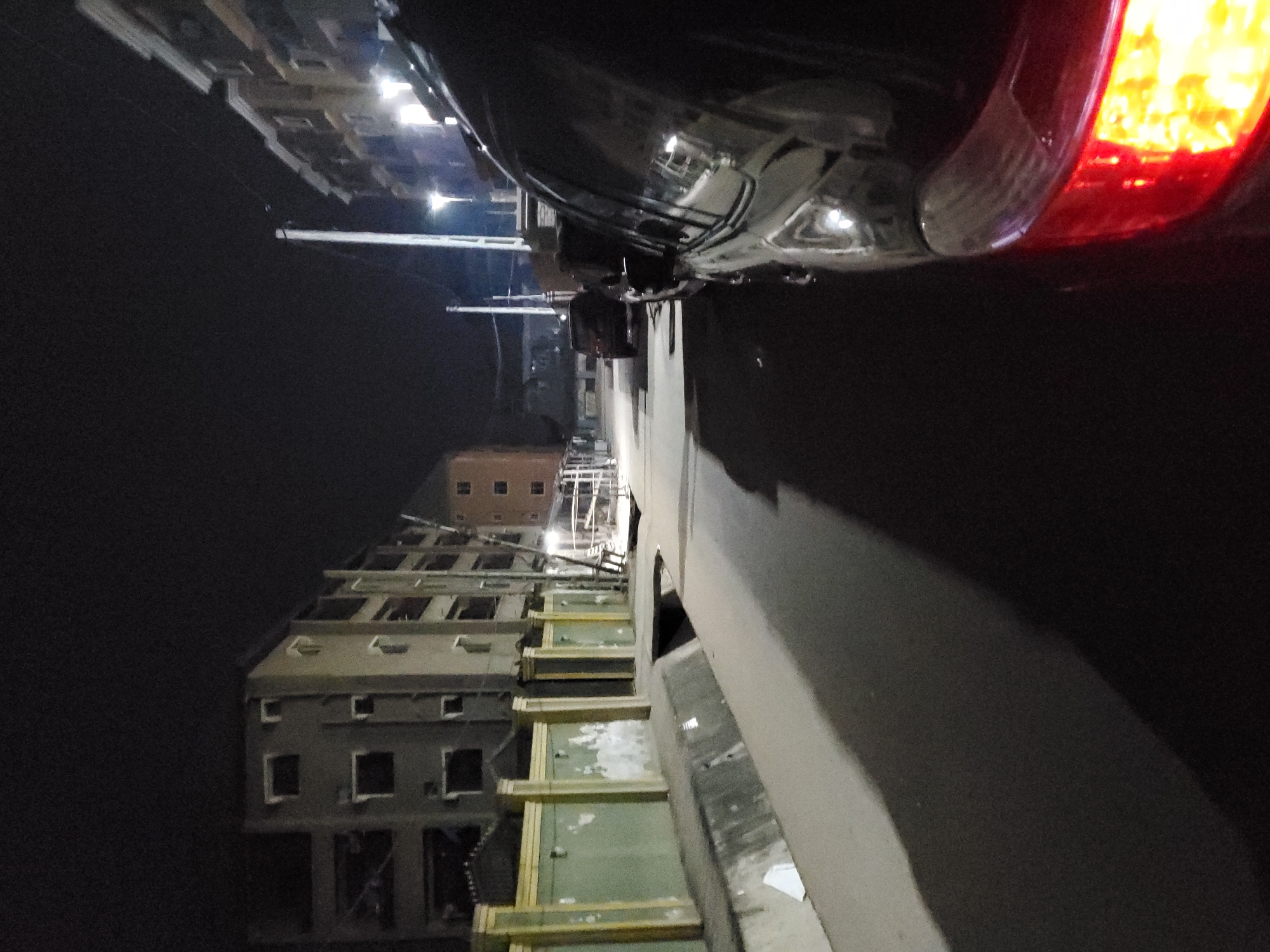 This nocturnal photograph, captured in Nkpor, Nigeria, depicts an urban street scene with a distinct vertical orientation. The image is composed of a dark, glossy vehicle in the foreground on the right, and a row of multi-story buildings stretching into the mid-ground on the left.
The foreground is dominated by the rear section of a dark-colored car, possibly black or dark blue, with reflections of distant lights on its shiny surface. Its bright red and orange taillight is illuminated, suggesting it might be stationary or braking.
To the left, a series of residential or commercial buildings rise vertically, appearing somewhat aged or in a state of disrepair. They feature numerous rectangular windows, some of which are dark voids, while others show glimpses of internal light or reflections. Portions of these buildings, particularly on lower levels, display faded greenish-yellow paint and what appears to be peeling plaster or exposed framework. Between the car and the buildings, in the middle distance, there's a complex structure that resembles scaffolding or the exposed skeleton of a building, with bright lights visible within it, possibly indicating ongoing construction or a light source from within a damaged structure.
The background shows further dimly lit buildings and a few distant streetlights or utility poles, extending into the darkness of the night. The overall lighting is low, with artificial light sources providing the main illumination, highlighting specific elements and casting deep shadows.
There are no people visible in the image, and no specific activity is taking place, giving the scene a still and quiet atmosphere characteristic of late-night hours. No discernible text is clearly visible on any signs or objects. The environment appears utilitarian, with the contrast between the dark night sky, artificial lights, and the rugged textures of the buildings and the car.
