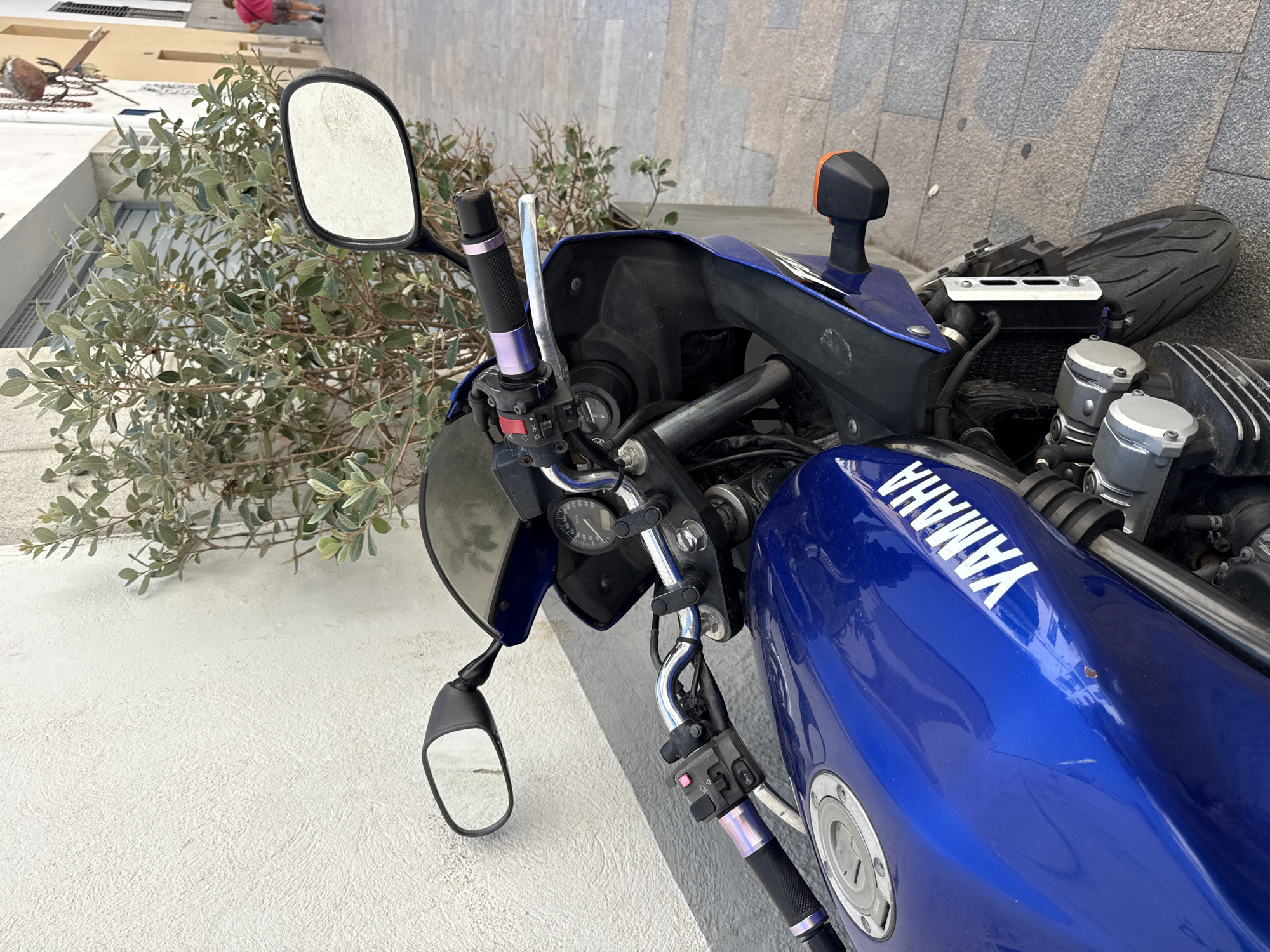 A blue Yamaha motorcycle is parked outdoors on a stone-tiled sidewalk next to a textured white wall. A green leafy bush grows against the wall behind the motorcycle. The motorcycle's handlebars are visible, along with a side mirror, part of the fuel tank with the "YAMAHA" logo, and the front wheel. In the background, a person in a pink shirt and shorts is walking away from the camera down the sidewalk. The street is lined with buildings and a hanging decorative item is visible on the side of one building.