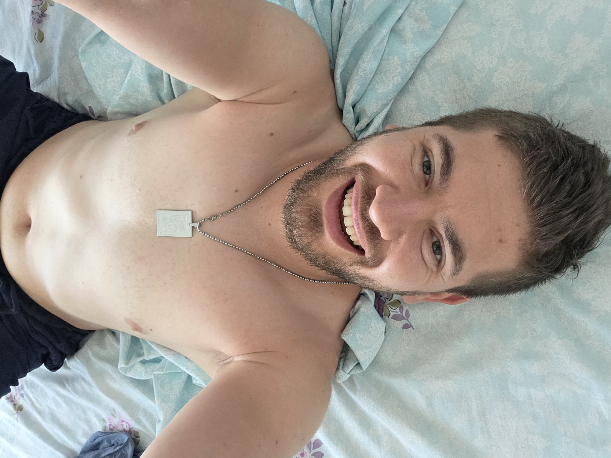 An overhead image depicts a male individual, appearing to be in his 20s or 30s, lying supine on a bed in Yagodovo, Bulgaria. He is bare-chested, with dark shorts or underwear visible at the top of the frame, partially covering his lower abdomen. His head is positioned at the lower edge of the frame, and he is looking directly at the camera with a smiling expression, mouth slightly open to reveal his upper teeth. He has short brown hair and a neatly trimmed brown beard. A silver-colored chain necklace with a rectangular, light-colored pendant displaying an embossed pattern rests on his chest. His arms are raised overhead, with hands out of frame, suggesting a selfie capture. The bed features light blue bedding; a fitted sheet has a subtle white pattern, while a crumpled top sheet exhibits a floral design of purple flowers and green leaves.