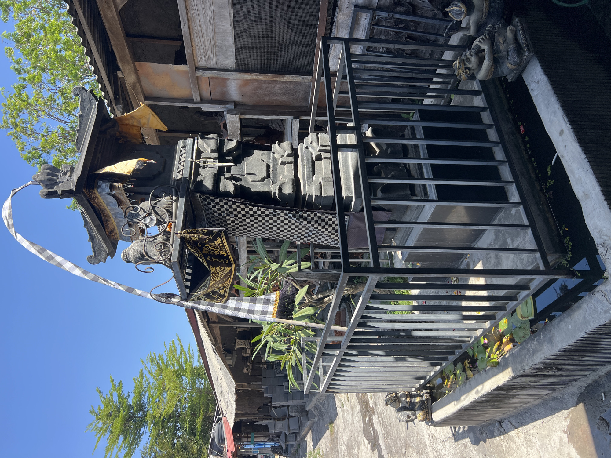 This is a medium shot, eye-level, exterior, daytime photograph of a shrine in Denpasar, Indonesia. The shrine is made of dark stone and features intricate carvings. It is decorated with traditional Balinese fabrics, including a black and white checkered cloth and a gold and black patterned cloth. A tall, slender flag, also black and white checkered, is attached to a pole and extends upwards from the shrine. The shrine is surrounded by a black metal fence, and in front of the fence is a narrow water feature containing lily pads. Two small, seated stone figures are positioned on the edge of the water feature. Behind the shrine, there are several buildings with corrugated metal roofs. Some of these buildings appear to be workshops or shops, with stacks of stone objects visible. A lush green tree is present in the background to the left, and the sky is a clear, bright blue. The overall impression is one of a sacred or culturally significant space within a bustling marketplace or craft area.