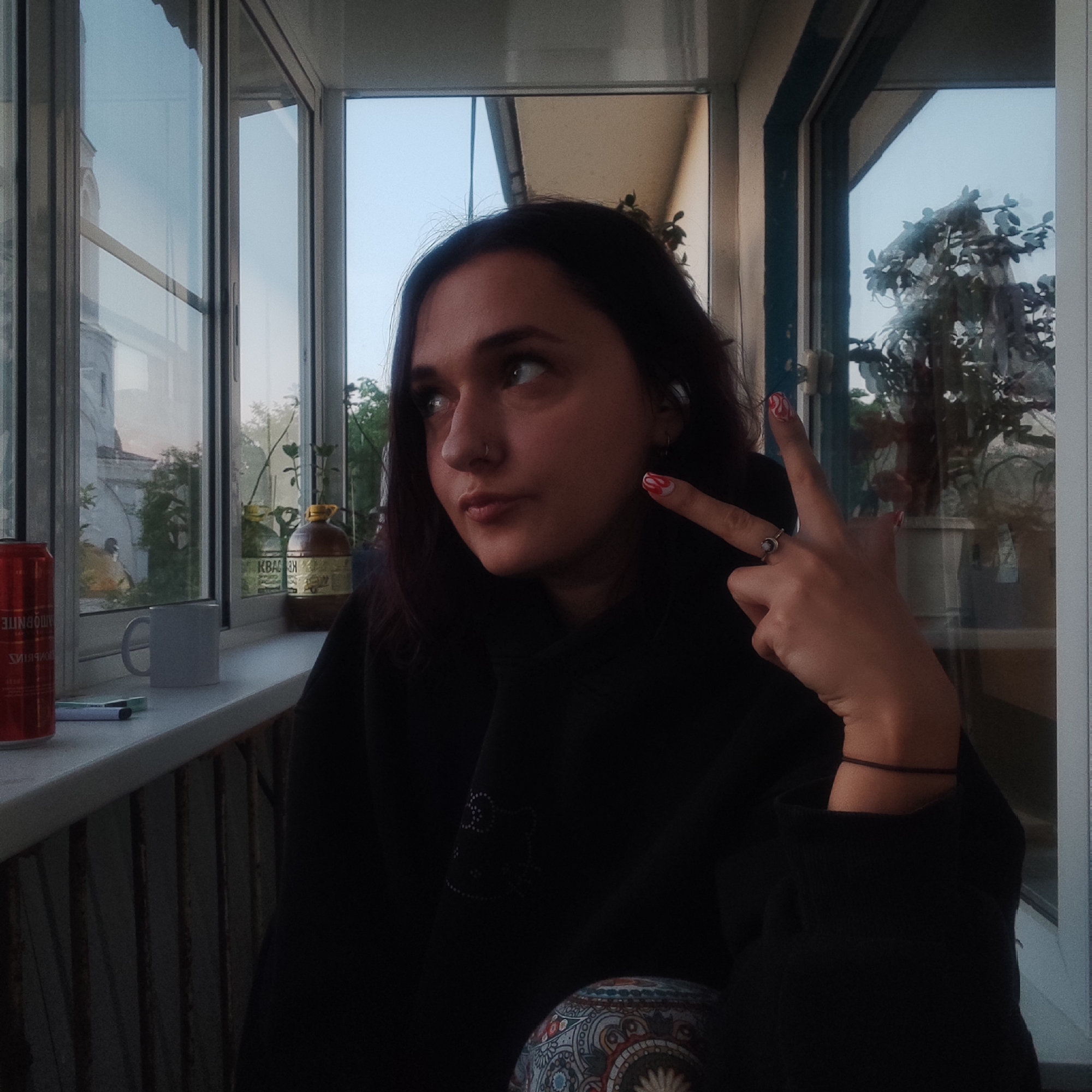 A young woman with dark hair and a nose piercing is seated in an enclosed balcony or loggia, possibly in Yekaterinburg, Russia. She wears a dark hooded top and colorful, patterned pants. Her gaze is directed upwards and to her left, with a pensive or slightly smirking expression. Her right hand is raised, making a "peace sign" with her index and middle fingers, and a distinct red and white swirl design is visible on her index fingernail. She also wears a ring on her ring finger.
The setting features large windows, reflecting an urban exterior with an older building (possibly a church or historic structure with an arched window) and trees. The light suggests late afternoon or early evening. On the windowsill, several items are placed: a red aluminum beverage can, a white ceramic mug, two markers (one blue, one green), and a brown plastic bottle with a yellow cap, clearly labeled "КВАС" (Kvas) in Cyrillic. Additional potted plants are visible both inside the loggia and reflected in the windows. The scene captures a quiet moment within an urban environment.