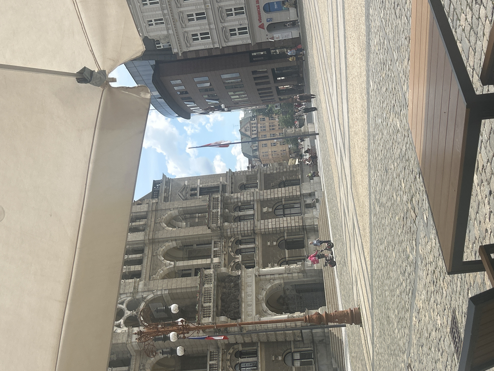 This urban scene, captured in Liberec, Czechia, portrays a lively city square under bright daylight conditions, with a mix of historical and modern architecture. The sky is partly cloudy with patches of blue.
In the immediate foreground, a large, light-beige outdoor umbrella or awning partially frames the left side of the image, while a portion of a wooden table with a dark metal frame is visible on the far right, suggesting an outdoor cafe. The ground is paved with a combination of grey cobblestones and smoother, striped light-colored flagstones.
The central feature of the image is a grand, intricately detailed stone building with a light grey facade, adorned with numerous arched windows, decorative cornices, and sculptural elements. In front of this building stands an ornate street lamp post with multiple lights. Further in the background, other buildings of varying architectural styles are visible, including one with modern glass elements, and a flagpole flying a red and white flag.
Several people are scattered throughout the square. Two individuals near the ornate building are riding personal transporters, possibly Segways. Other people are seen walking or standing, contributing to the active atmosphere of the square. On a building in the mid-distance, a blue sign with white text is visible, clearly displaying "VZ VESMÍR" and "ODĚVY", indicating a business. The scene conveys a calm and ordinary day in a European city.
