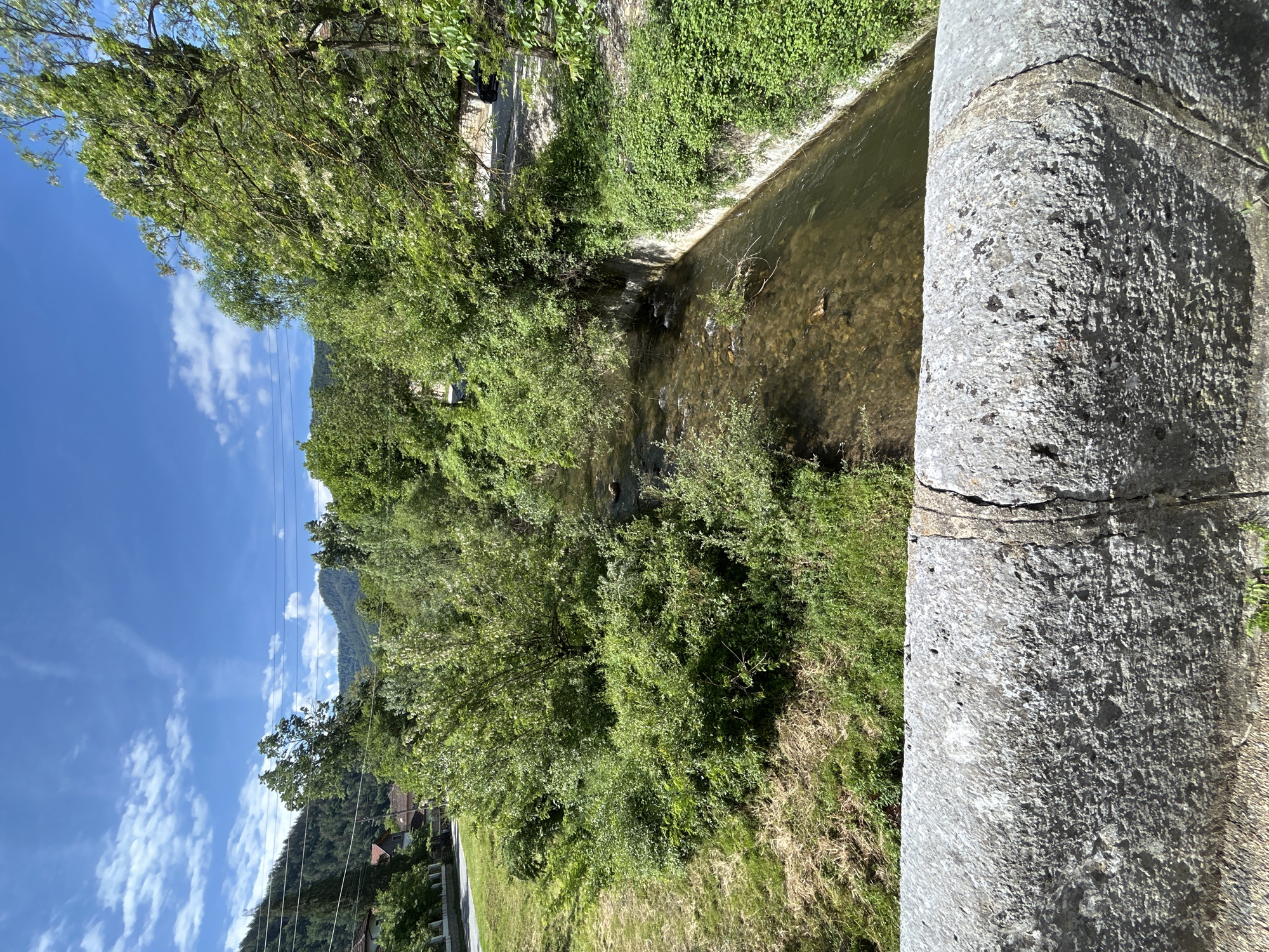 The image presents a vibrant outdoor scene centered around a narrow stream or river. A prominent, rough-textured concrete structure, likely a bridge pillar or railing, occupies the right foreground, partly obscuring the view of the water directly beneath.
The stream flows through the mid-ground, its banks densely lined with lush green trees and bushes. The water appears clear, revealing the rocky riverbed below, with small ripples indicating a gentle current. The vegetation on both sides of the stream is verdant, showcasing various shades of green, suggesting a healthy ecosystem. Some trees have lighter green or whitish foliage, possibly new spring growth or blossoms.
Beyond the immediate riparian zone, in the background, a range of green, forested mountains rises under a bright blue sky. Scattered white cumulus clouds are visible against the clear sky, indicating sunny weather. Multiple parallel power lines stretch horizontally across the mid-ground, spanning the distance between the trees and the mountains. Hints of human habitation, such as parts of buildings with reddish roofs, can be discerned nestled among the trees near the base of the mountains.
The time of day is clearly daylight, likely mid-day, given the bright illumination and distinct shadows. No specific activity or people are visible; the scene is a static capture of a natural and semi-rural landscape. The concrete structure in the foreground exhibits a clear horizontal crack. No text is visible in the image.