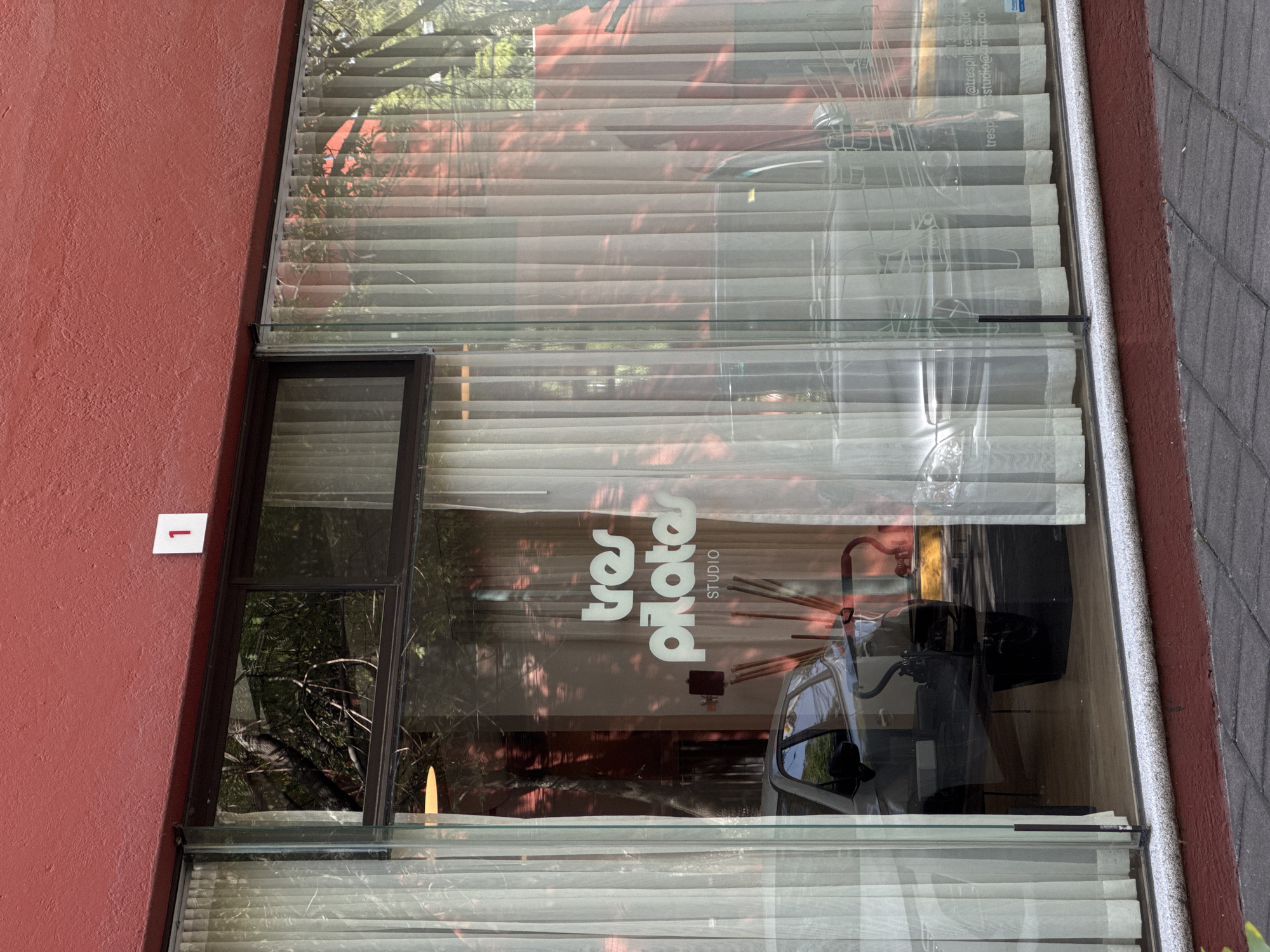 The image captures the exterior of a business in Puebla, Mexico, identified as "tres pilates STUDIO," as indicated by the logo displayed on the window. The building's exterior is painted in a reddish-brown hue, with the number "1" displayed on a white square next to the business. The window is covered with closed blinds and sheer white curtains, partly obscuring the view inside. Reflections on the glass show glimpses of the surrounding environment, including trees and shadows. The presence of pilates equipment can be seen in the background through the window. The building abuts a gray-paved sidewalk.