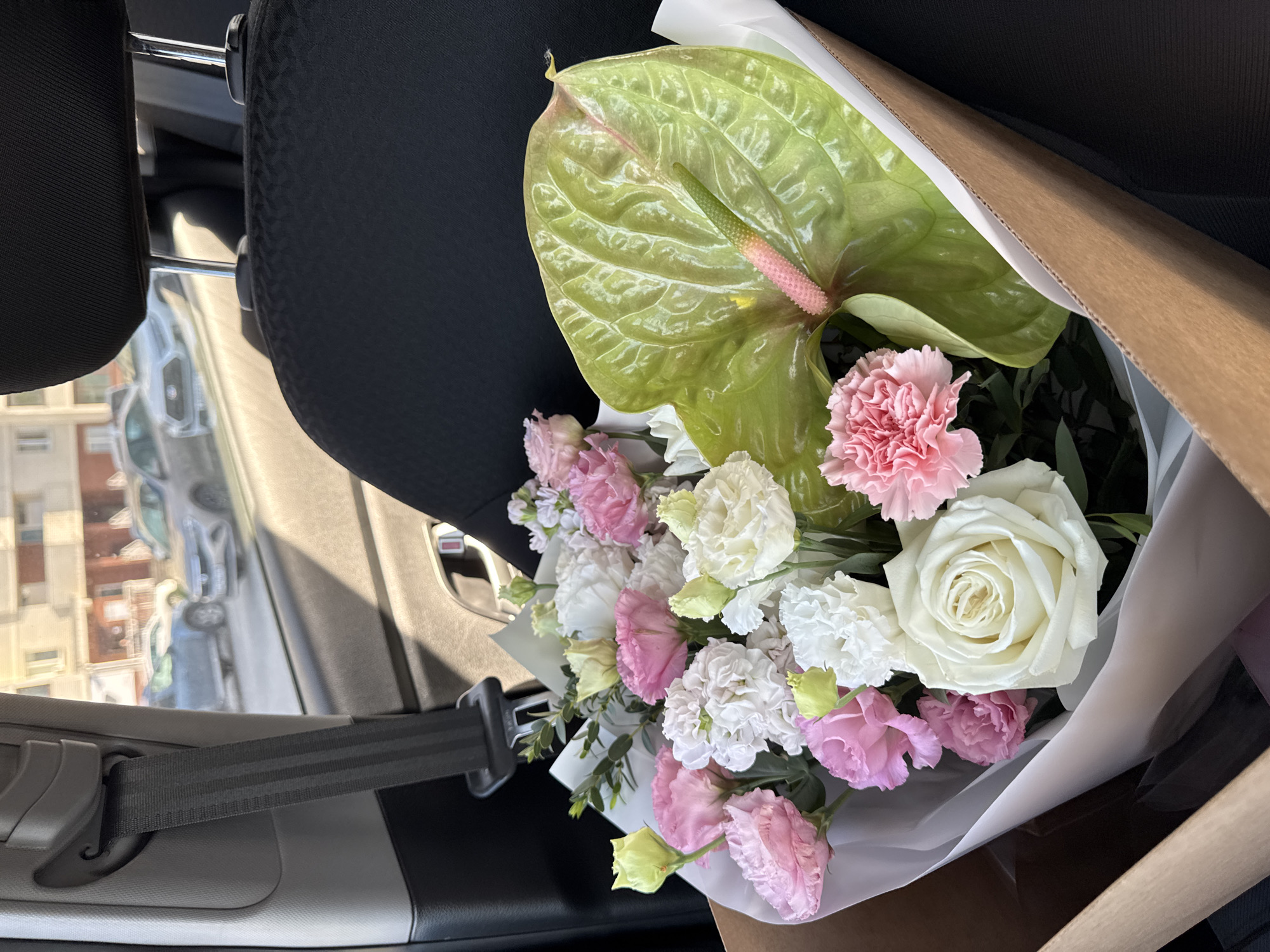 A large bouquet of fresh flowers, wrapped in white tissue paper and a brown cardboard holder, occupies the foreground, positioned within the interior of a vehicle. The bouquet features a variety of blooms, including white roses, pink and white Eustoma (Lisianthus), pink carnations, and a prominent green Anthurium spathe with a distinct pink spadix. The car's interior is visible, showing a black fabric seat and a grey seatbelt mechanism on the left. Through the car window in the background, an urban environment in Izhevsk, Russia, is depicted under natural daylight. Multi-story, light-colored residential buildings line a street where several cars are parked or in motion, including a light-colored SUV on the right and a darker vehicle further back on the left. No individuals are visible, and no specific actions or interactions are occurring.