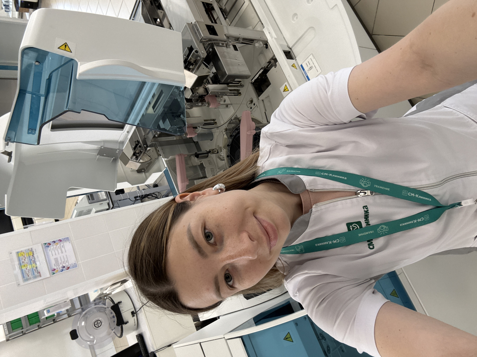 A young woman with light brown hair pulled back is taking a selfie in what appears to be a laboratory or medical testing facility. She is wearing a white lab coat with a green lanyard around her neck that has the logo "СМ-КЛИНИКА" printed on it. This logo indicates she is likely an employee of a medical clinic chain in Russia, as "СМ-КЛИНИКА" is a known private medical network. The background shows sophisticated medical equipment, including a large automated analyzer with a transparent blue cover. In the background on the left, a smaller piece of equipment that resembles a centrifuge is visible. There are also some charts and notices on the wall behind her. The overall environment suggests a professional medical setting. The specific location in Moscow is not explicitly provided by visual cues in the image.