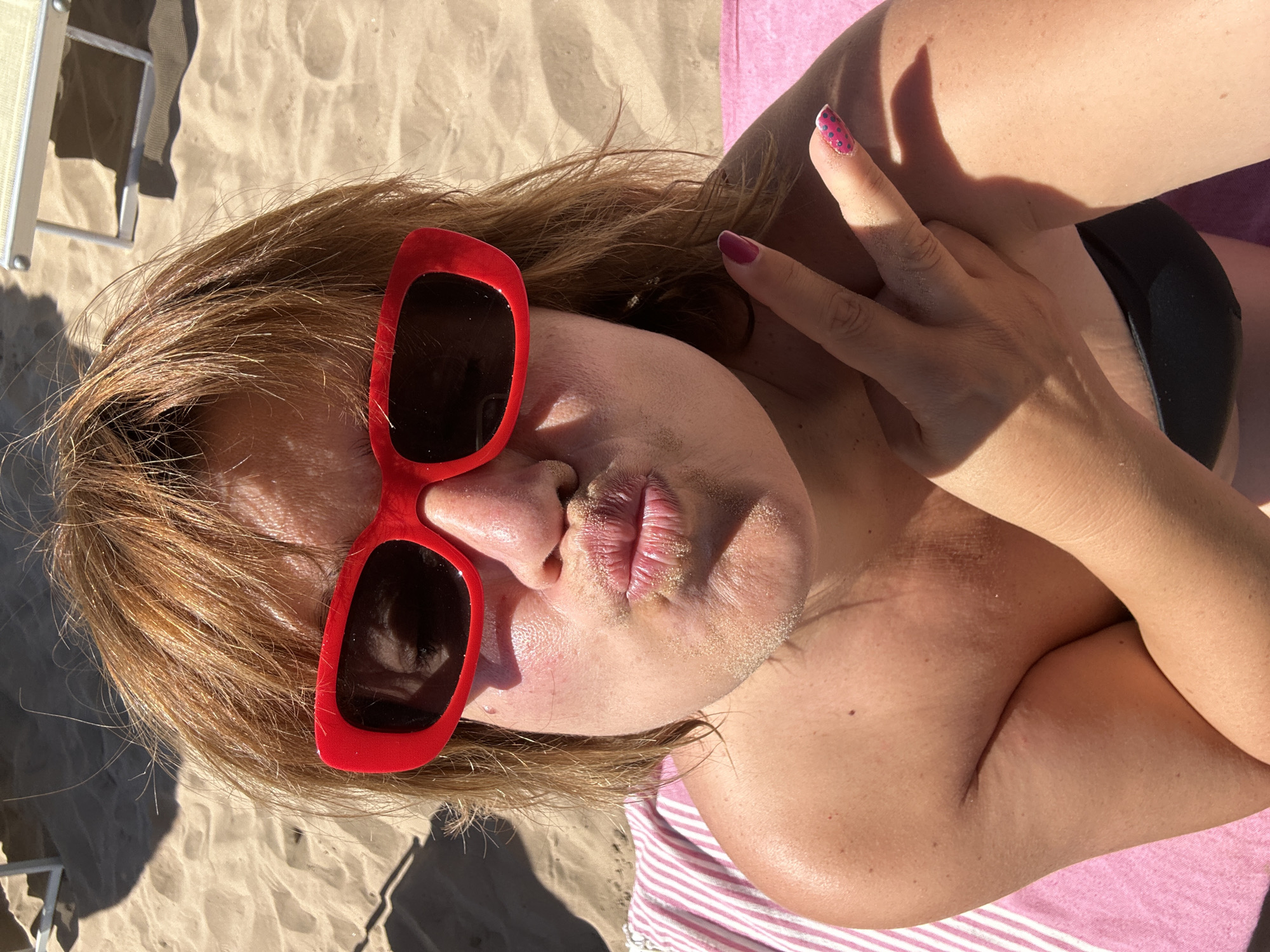 A medium shot captures a woman with auburn hair and red sunglasses posing for a selfie on a sandy beach. Her lips are puckered, with sand adhering to them. She is wearing a black bikini top and has her right hand raised in a peace sign, with fingernails painted pink and adorned with black polka dots. A pink and white striped towel is visible beneath her left shoulder, and a lounge chair casts a shadow in the sandy background. The scene appears to be in Peschici, Italy, given the context of the request.