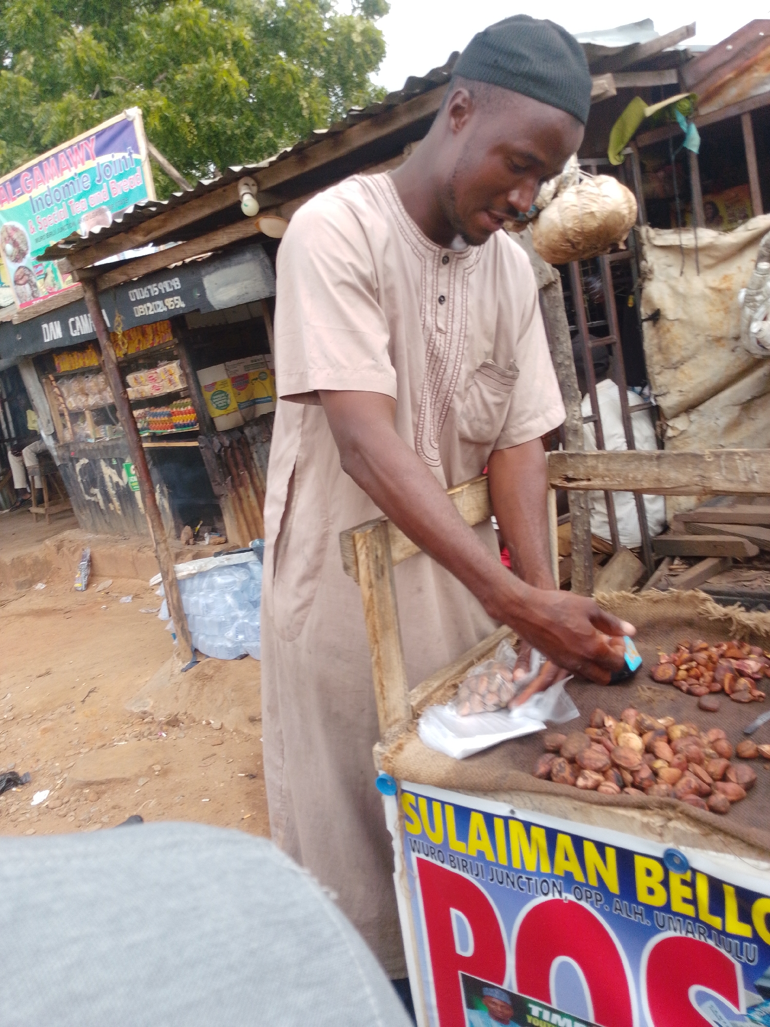The image shows a street vendor in Gombe, Nigeria, arranging nuts on a display table. The vendor, a man wearing a tan kaftan and a black cap, is focused on his task, his hands hovering over the nuts. The table is laden with a variety of nuts, some in small piles, others scattered. A sign on the table reads "SULAIMAN BELLO" in large yellow letters, with additional text below indicating "WURO BIRJI JUNCTION, OPP. ALH. UMAR LULU." The background reveals a bustling marketplace with makeshift stalls, corrugated metal roofing, and stacks of goods. Trees are visible in the distance, suggesting an outdoor setting. The ground is sandy and uneven, typical of many market areas in the region. The overall impression is one of everyday commerce and activity in a Nigerian city.