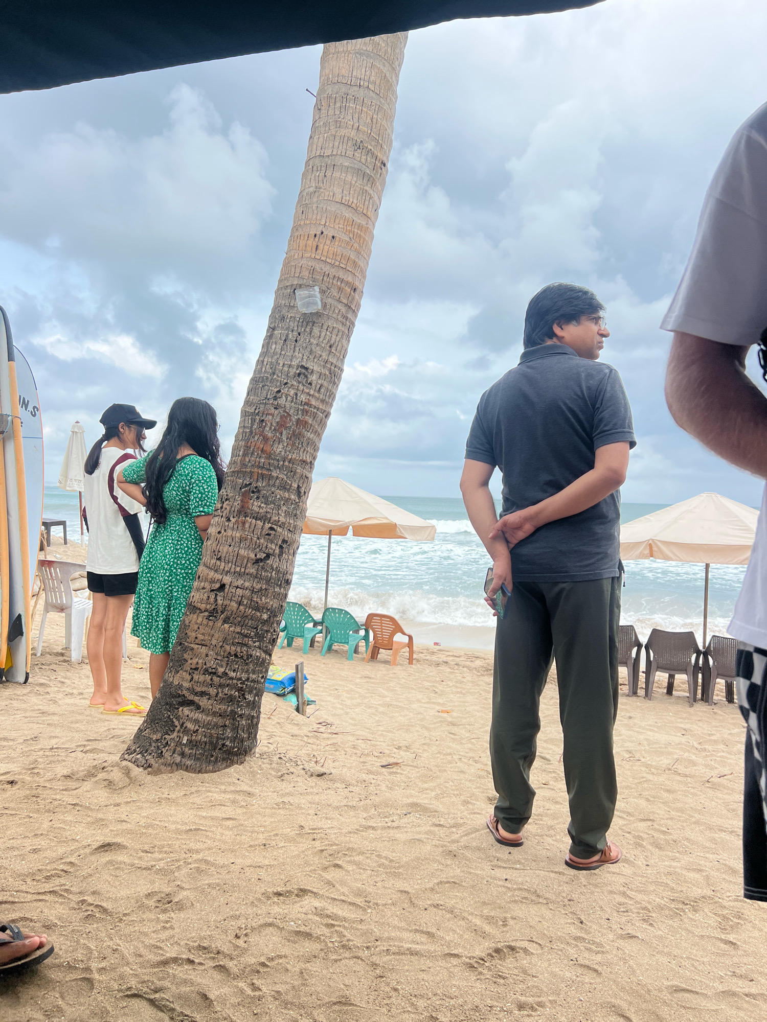 This image captures a scene at Kuta Beach in Kuta, Indonesia, on an overcast day. The foreground is dominated by sand and the base of a large palm tree with a visible plastic cup taped to its trunk.
In the mid-ground, a man in a dark grey polo shirt and dark green pants stands facing away from the camera, with his hands clasped behind his back. He is wearing brown sandals. To his left, two women are standing near the palm tree. The woman on the right wears a green floral dress, and the woman on the left wears a white t-shirt with a red stripe and black shorts, and a black baseball cap. Surfboards are visible to the far left of the frame.
In the background, the ocean is visible with moderate waves. Several beach chairs and umbrellas are set up on the sand. The sky is cloudy, contributing to the diffused lighting of the scene. The overall impression is a relaxed, outdoor setting at a popular beach destination.