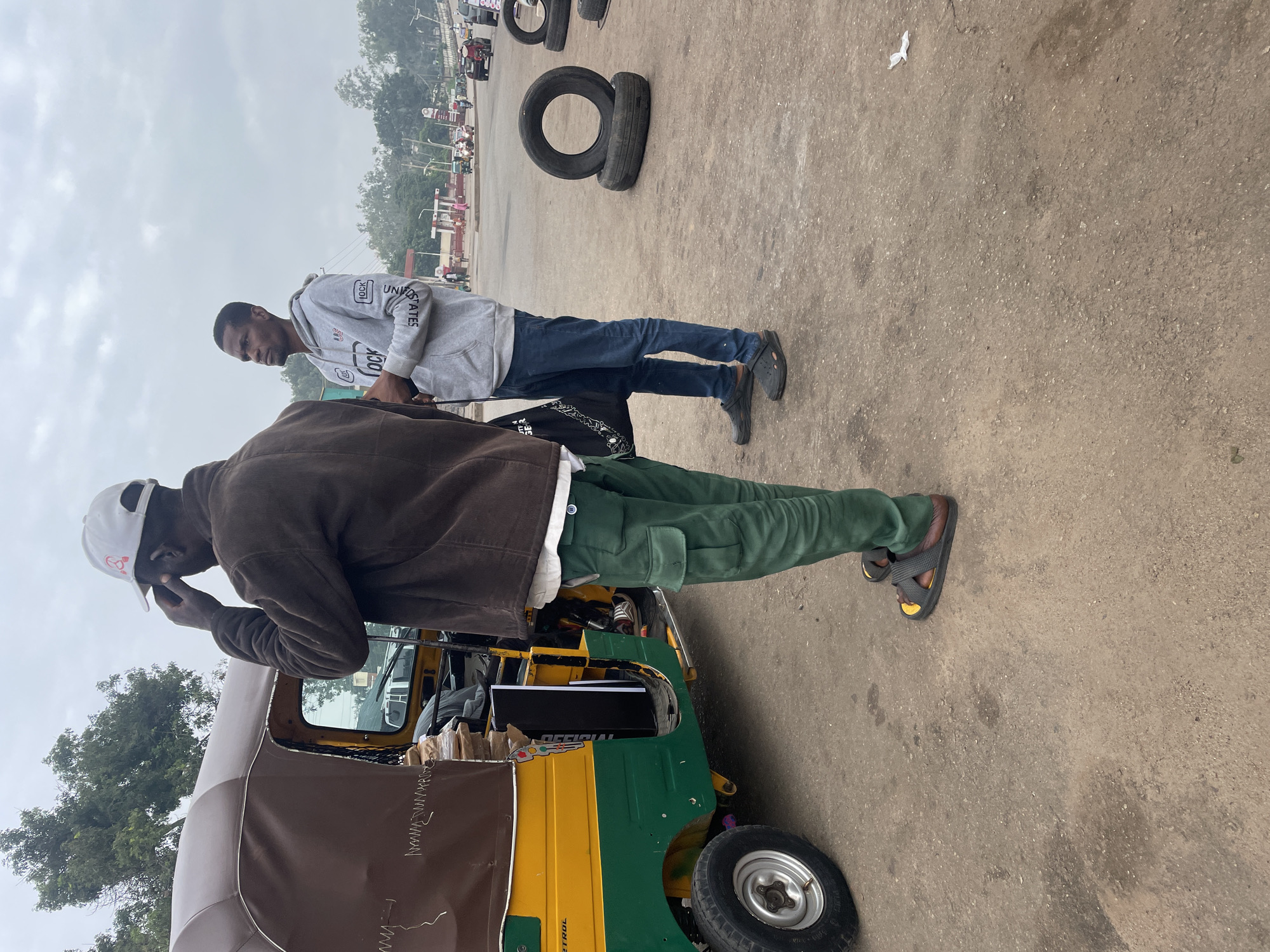 Two men stand near a three-wheeled vehicle, likely a tuk-tuk, on a dirt road in Jos, Nigeria. The man on the left, wearing a brown jacket and green cargo pants, is facing away from the camera, adjusting his white baseball cap. The man on the right, wearing a gray hoodie and blue jeans, stands with his hands in his pockets, looking towards the distance. Several tires are stacked on the ground to the right of the men. In the background, gas stations, buildings, and other vehicles suggest a busy urban environment. The sky is overcast, indicating a cloudy day. The overall scene depicts a street corner in Jos, possibly a transportation hub or market area.