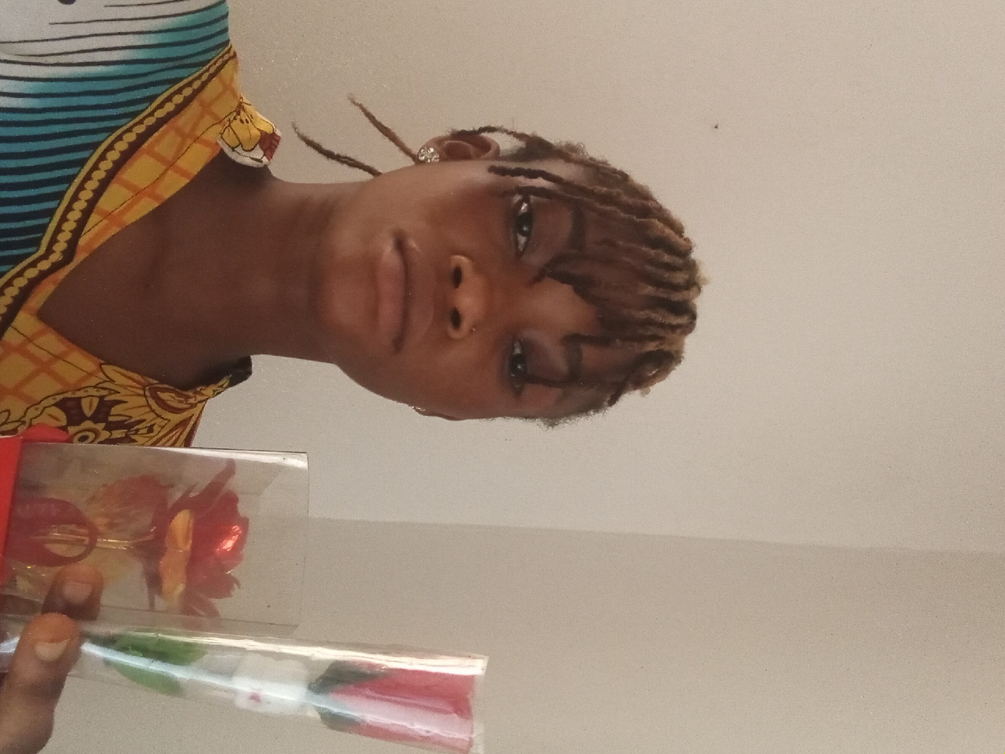 A young, Black woman with brown, braided hair poses, facing slightly left of the camera. She is holding two encased roses in her left hand. The top rose is in a clear box with a red bow, while the other is encased in clear plastic. She wears a yellow and brown patterned top over a white top with teal stripes. The background is a plain, light brown wall. The location is Unknown, Unknown.