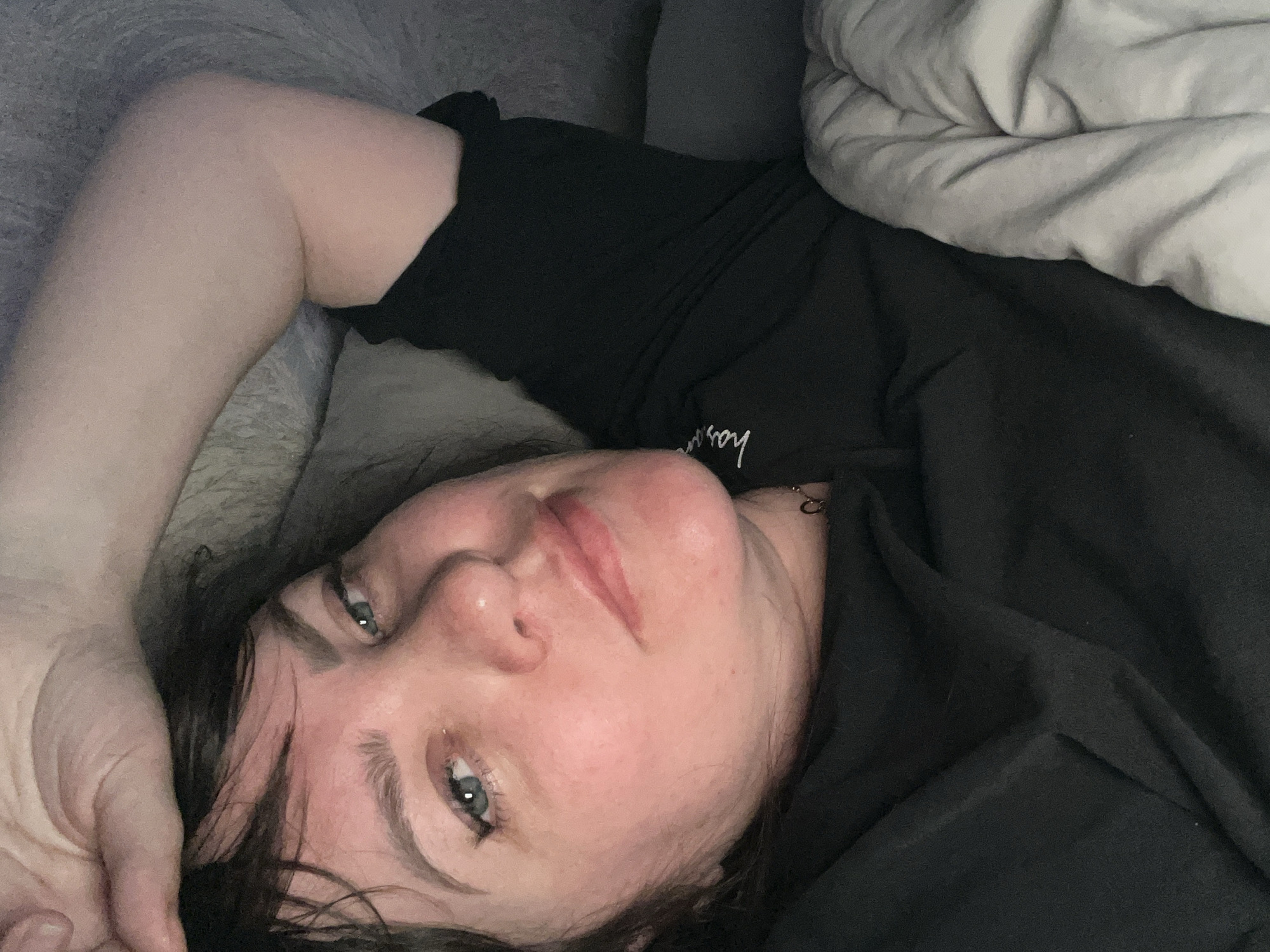A person is depicted lying on their back indoors, captured from an overhead, tilted perspective. The individual, with fair skin and dark, somewhat disheveled hair, has blue-green eyes that are open and directed towards the camera. A small piercing is visible on their left nostril. Their lips are slightly parted, revealing natural lip color.
The person is wearing a black short-sleeved top or t-shirt. Faint white script text, possibly reading "hosann" or similar, is partially visible on the lower right side of the garment. One arm is raised and rests on a soft grey surface, likely a pillow or cushion, which is positioned behind their head. A light grey or off-white duvet or blanket covers the lower part of their body, extending towards the top right of the frame.
The setting appears to be an indoor space, such as a bedroom or living room in Noviy, Russia, suggesting a moment of rest or relaxation. The lighting appears dim or artificial. The person's expression is neutral, possibly conveying a sense of calm, contemplation, or tiredness.