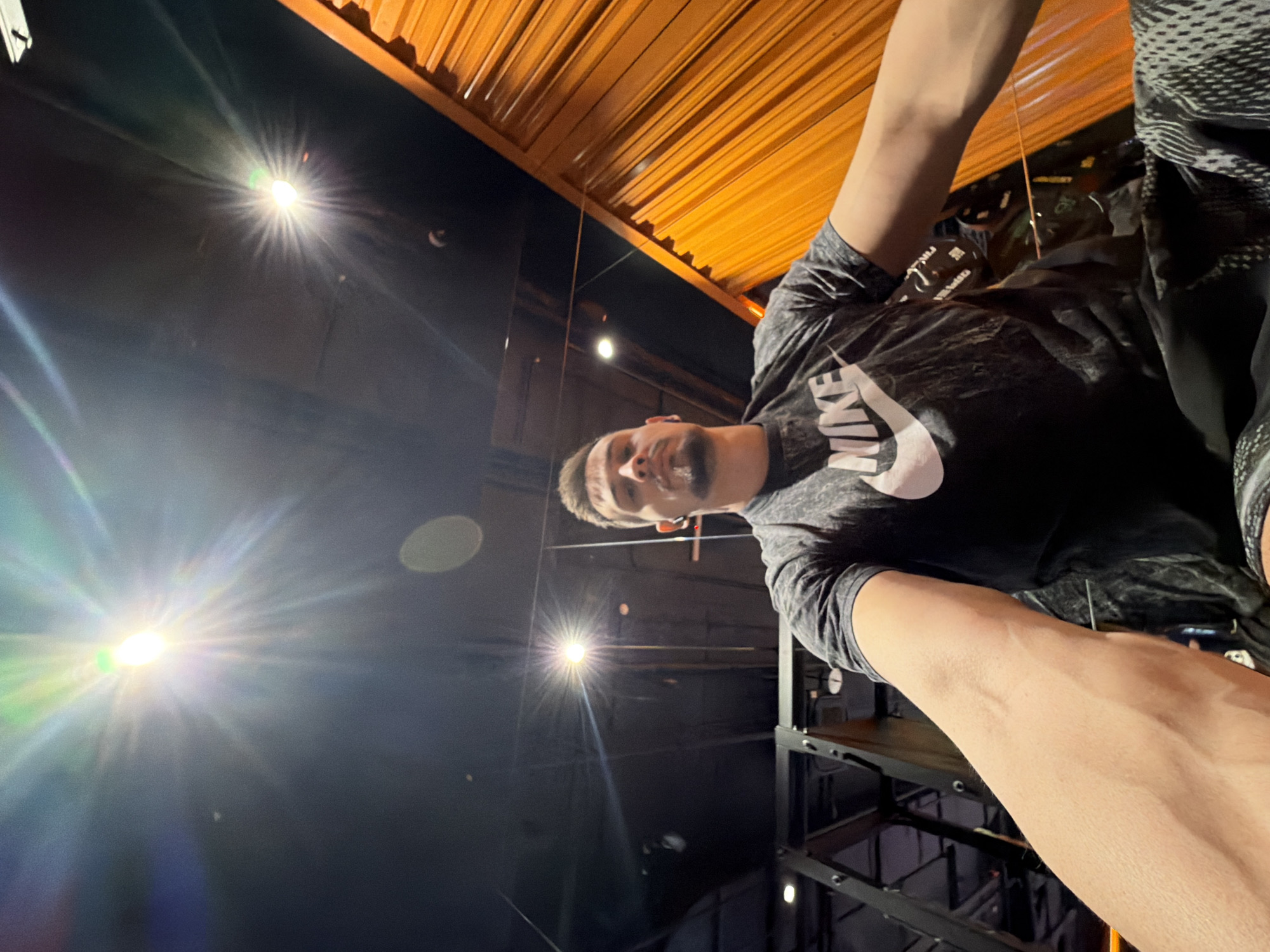 A low-angle selfie shows a man wearing a gray Nike t-shirt. The background features a black wall and an orange ceiling, along with bright lights. The location is the Grand Hall Siberia in Krasnoyarsk, Russia.