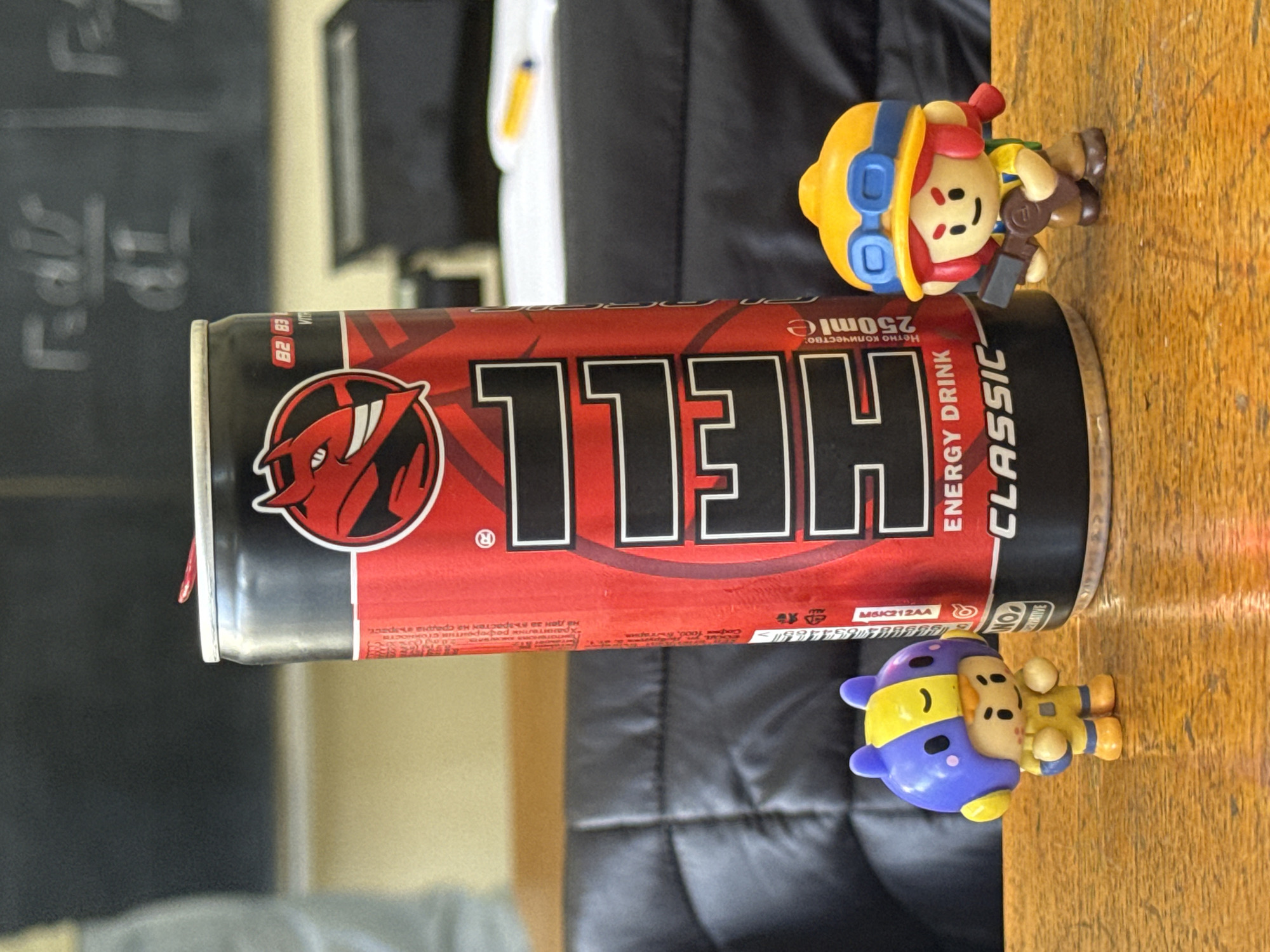 The image shows a 250ml can of "HELL Classic Energy Drink". The can is red with a black background, featuring the white "HELL" logo. A small, cartoon-style figurine of a person wearing a yellow helmet and blue goggles is positioned on top of the can. Another similar figurine is situated below the can. The can and figurines are resting on a wooden surface. In the background, there is a black leather surface. The text on the can includes "ENERGY DRINK" and "CLASSIC" along with other writing, including "София Кооб". The can also has "82 83 VITR" printed on it. The image doesn't provide enough information to determine if it was taken in Sliven, Bulgaria.
