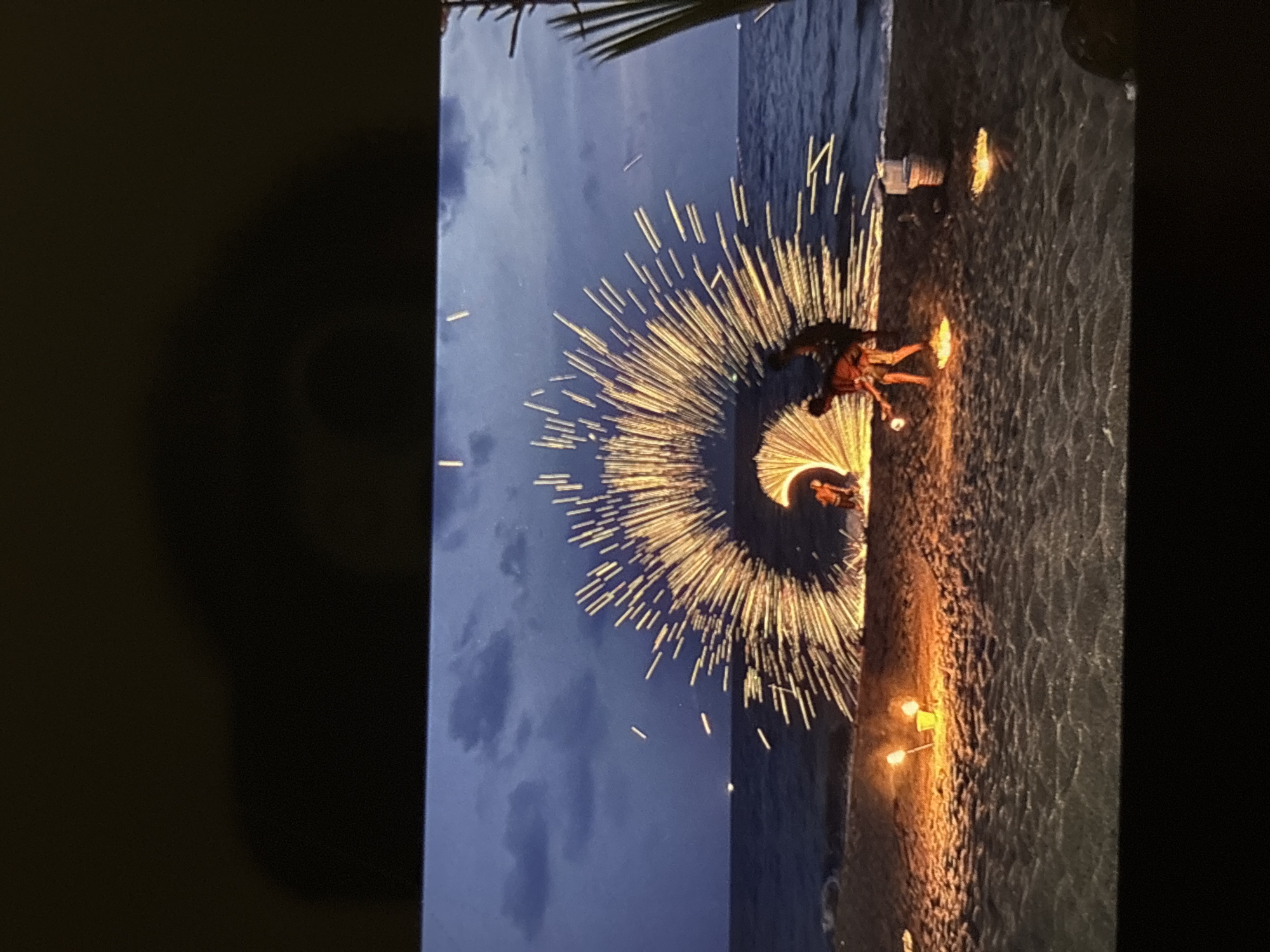 The image depicts a fire show performance on a beach at dusk. The performers are spinning fire, creating a large circular arc of sparks that extends upwards and outwards. Two individuals are visible in the foreground, one holding a fire prop and the other appearing to be the focus of the fire spinning. The beach is sandy, with the ocean visible in the background under a twilight sky with clouds. Several small fires or lights are visible on the sand, possibly marking the performance area. The scene is consistent with activities often seen on beaches in tourist areas like Chalong, Thailand.