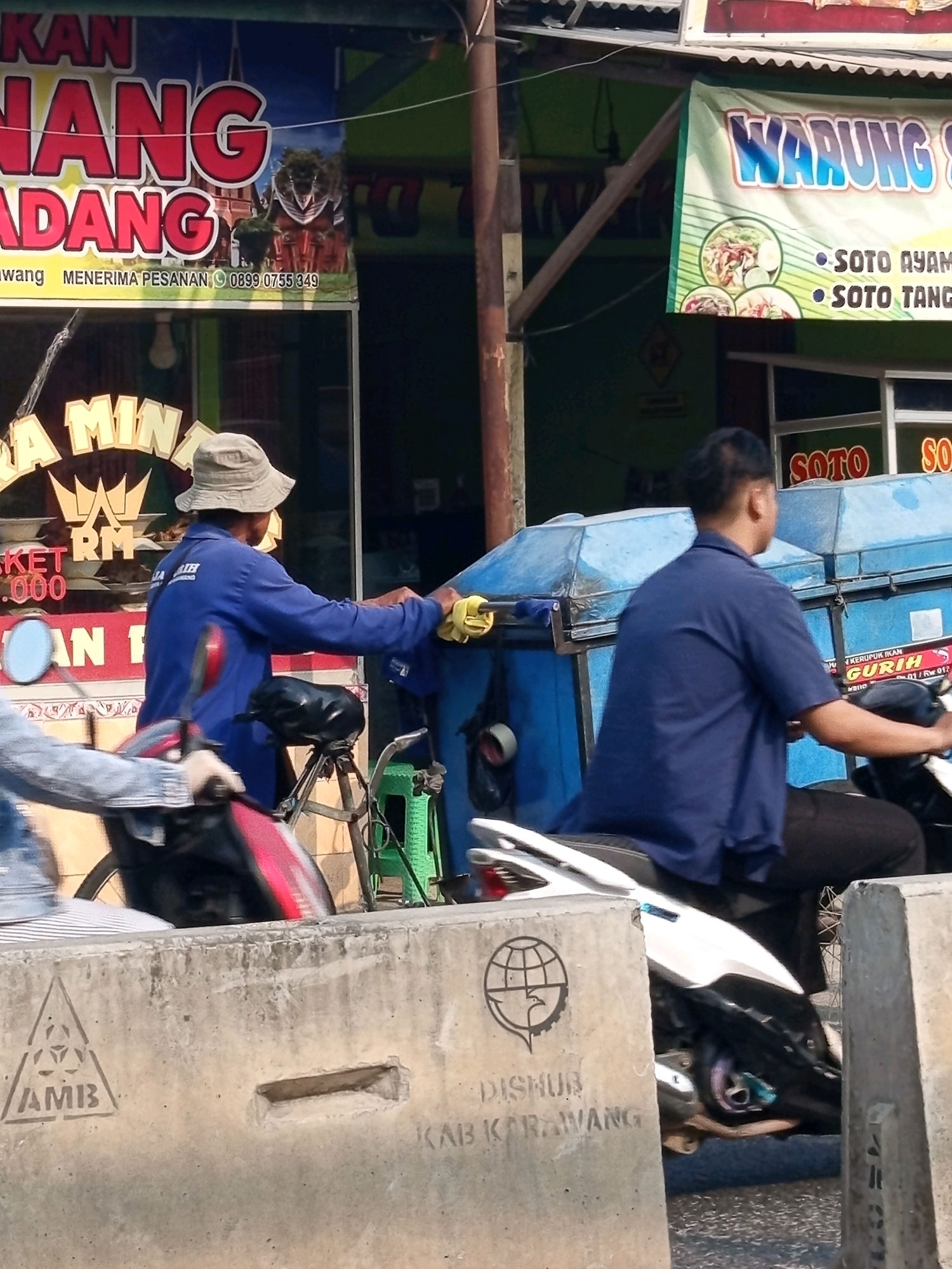 The image depicts a street scene in Karawang, Indonesia, characterized by commercial signage and street infrastructure. Dominating the background is a large banner advertising "KAN NANG PADANG" with phone number 0899 0755 349 and "MENERIMA PESANAN." To the right, a green banner reads "WARUNG SOTO" and lists menu items like "SOTO AYAM" and "SOTO TANG." In the mid-ground, a person wearing a blue long-sleeved shirt and black pants is seated on a white motorcycle. They are holding a yellow cloth, possibly cleaning or wiping something on a blue container attached to a cart or another vehicle. Another person in a denim jacket is partially visible on a red and black motorcycle in the foreground. The scene is bordered by concrete barriers in the foreground, one bearing a stylized globe and bird emblem above the stencil "DISHUB KAB KARAWANG." The overall environment suggests a busy commercial area with transportation elements.