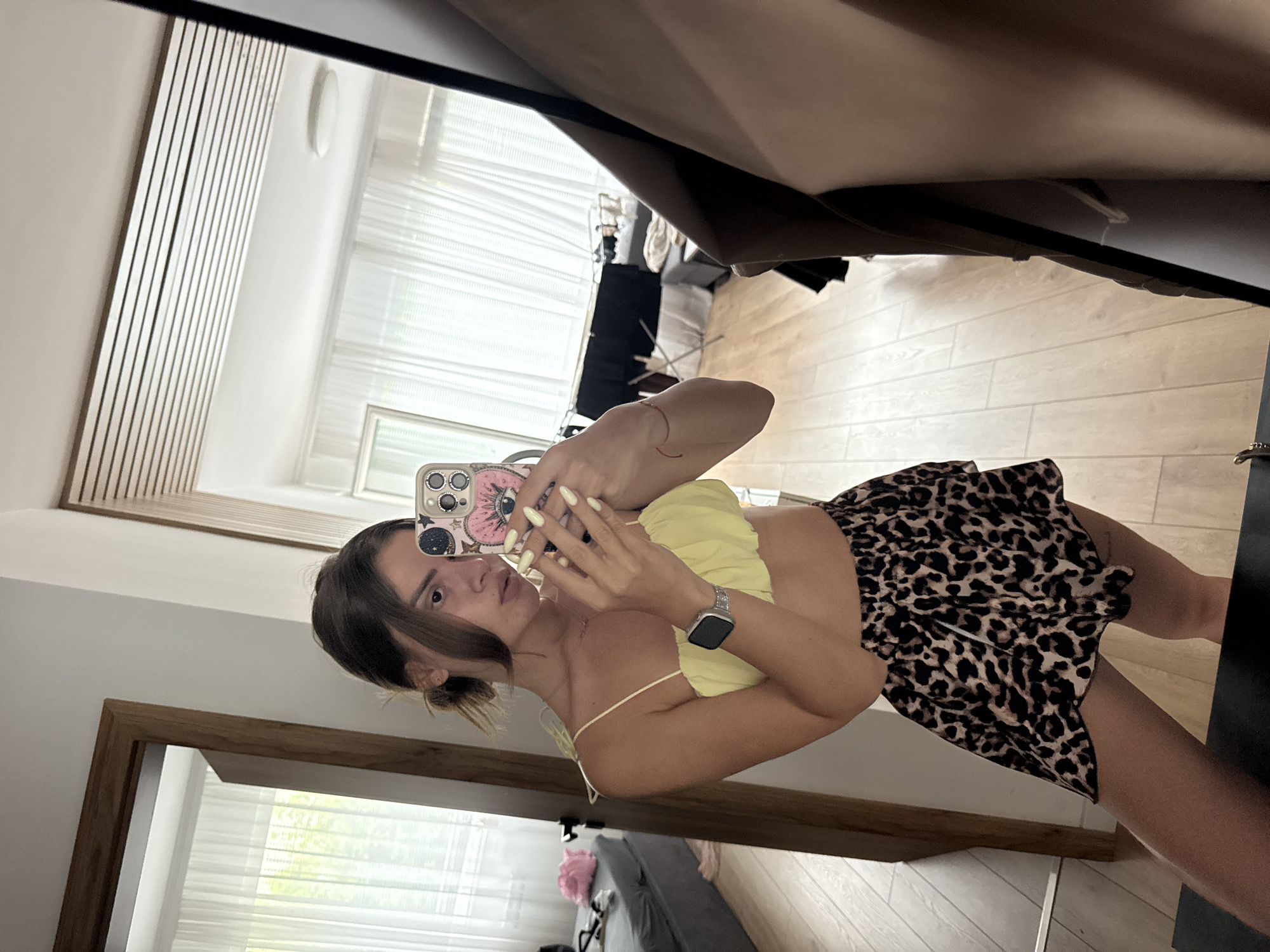 A young woman with dark hair pulled back takes a selfie in a mirror. She is wearing a yellow ruffled crop top and a leopard print mini skirt. A silver smartwatch is visible on her left wrist. The background shows a bedroom with a bed, a doorway, and a large mirror reflecting more of the room, including white curtains and a drying rack. The flooring is light-colored wood. The location is not specified as being in Plovdiv, Bulgaria.